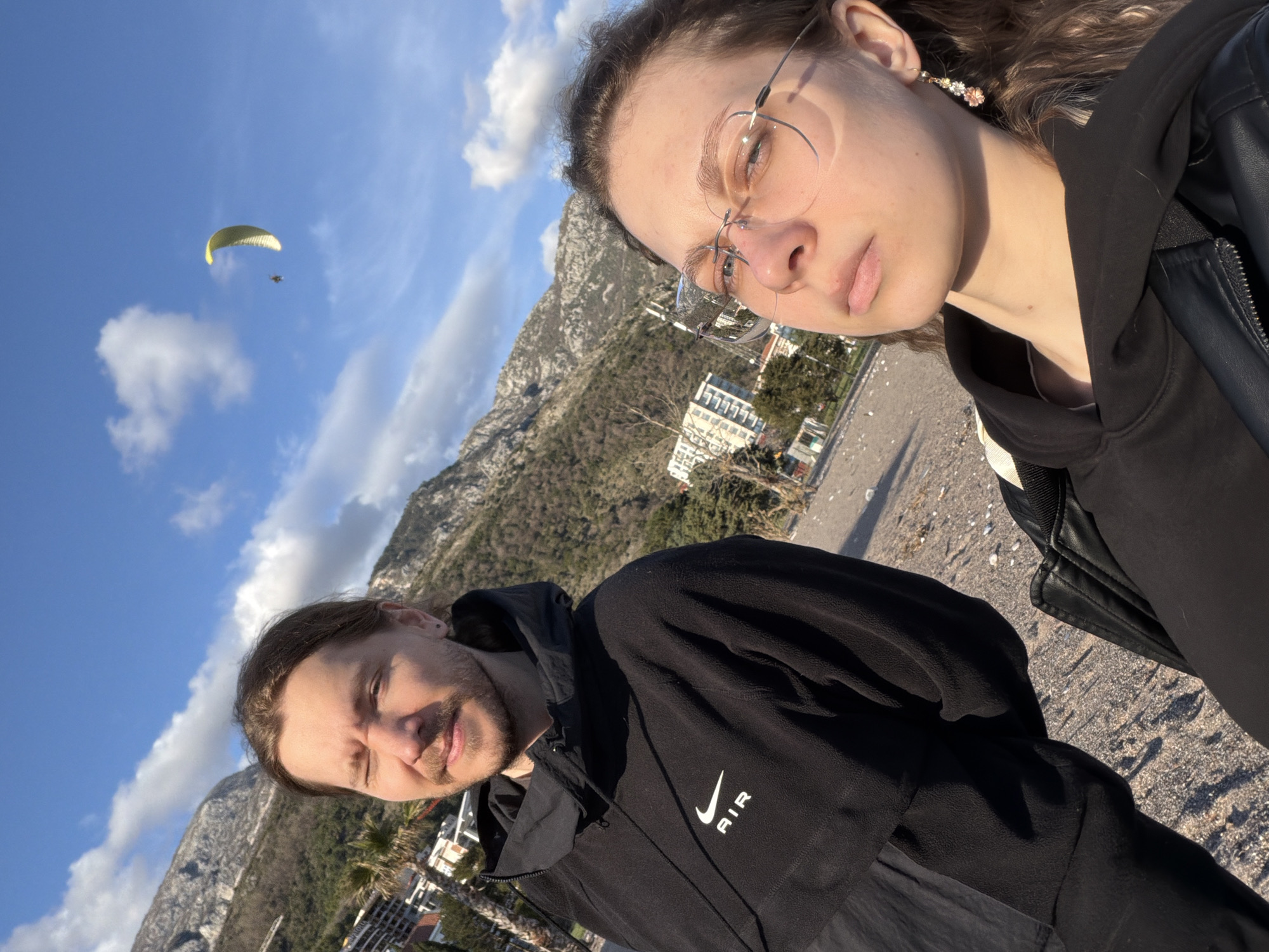 A selfie taken in Boreti, Montenegro, features two people. The person in the foreground on the right wears glasses and a black hooded jacket, with dangling floral earrings. The other person is positioned lower and to the left, wearing a black jacket with "AIR" and a Nike swoosh logo visible.
In the background, there's a mountainous terrain, buildings, and clear blue sky with scattered clouds. A yellow paraglide is visible in the upper left corner of the image. The ground appears to be a paved area.