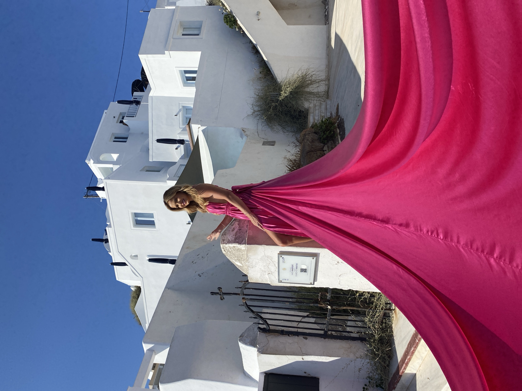A woman in a long, vibrant fuchsia dress with a thigh-high slit stands on a staircase in front of a white building with blue-domed roofs and dark umbrellas on the terraces. The dress trails behind her, creating a dramatic sweep across the foreground. The woman is smiling and looking to her left. The architecture is typical of the Cycladic islands, likely in Thira, Greece, known for its white-washed buildings and blue accents. The background shows more white buildings and a clear blue sky. A sign on a post next to a gate reads "CHURCH OF ANASTASI".