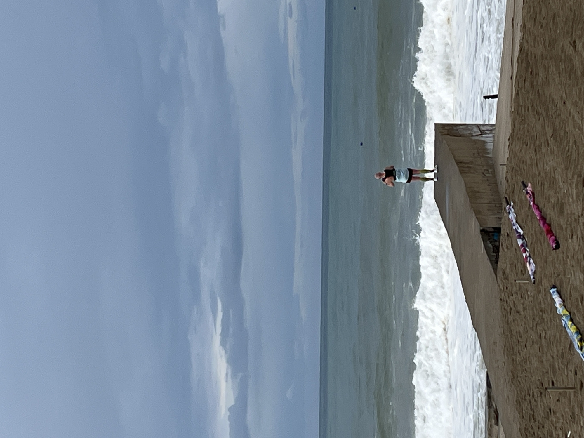 The image captures a coastal scene, likely in Alanya, Turkey, characterized by a pier extending into the sea. On the pier stands a lone person, potentially observing the waves crashing against the structure. The sky is overcast with thick clouds. The beach is visible. The presence of possible prayer flags is seen on the beach near the pier.