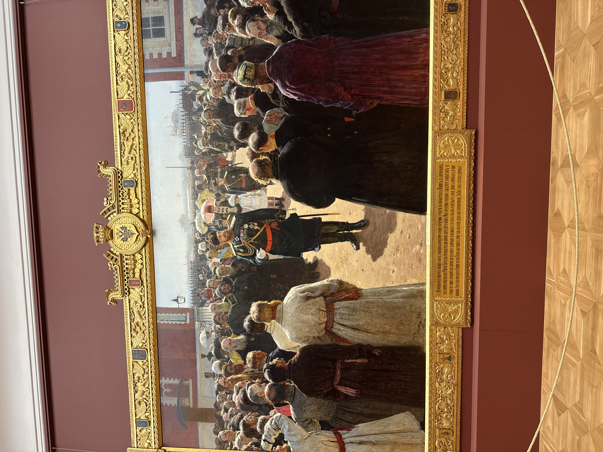 The image shows a large oil painting depicting a historical event. A crowd of people, many wearing dark clothing, are gathered in a square in Moscow, Russia. In the center of the crowd, a military figure in a green and black uniform is holding a speech. The painting is encased in an ornate, gilded frame. The location is likely in or near the Moscow Kremlin, given the presence of red brick buildings. The image also captures details of the room where the painting is displayed, including a deep red wall and a parquet floor. There's a rope barrier in the background. The painting includes many individual portraits within the crowd, creating a sense of realism.