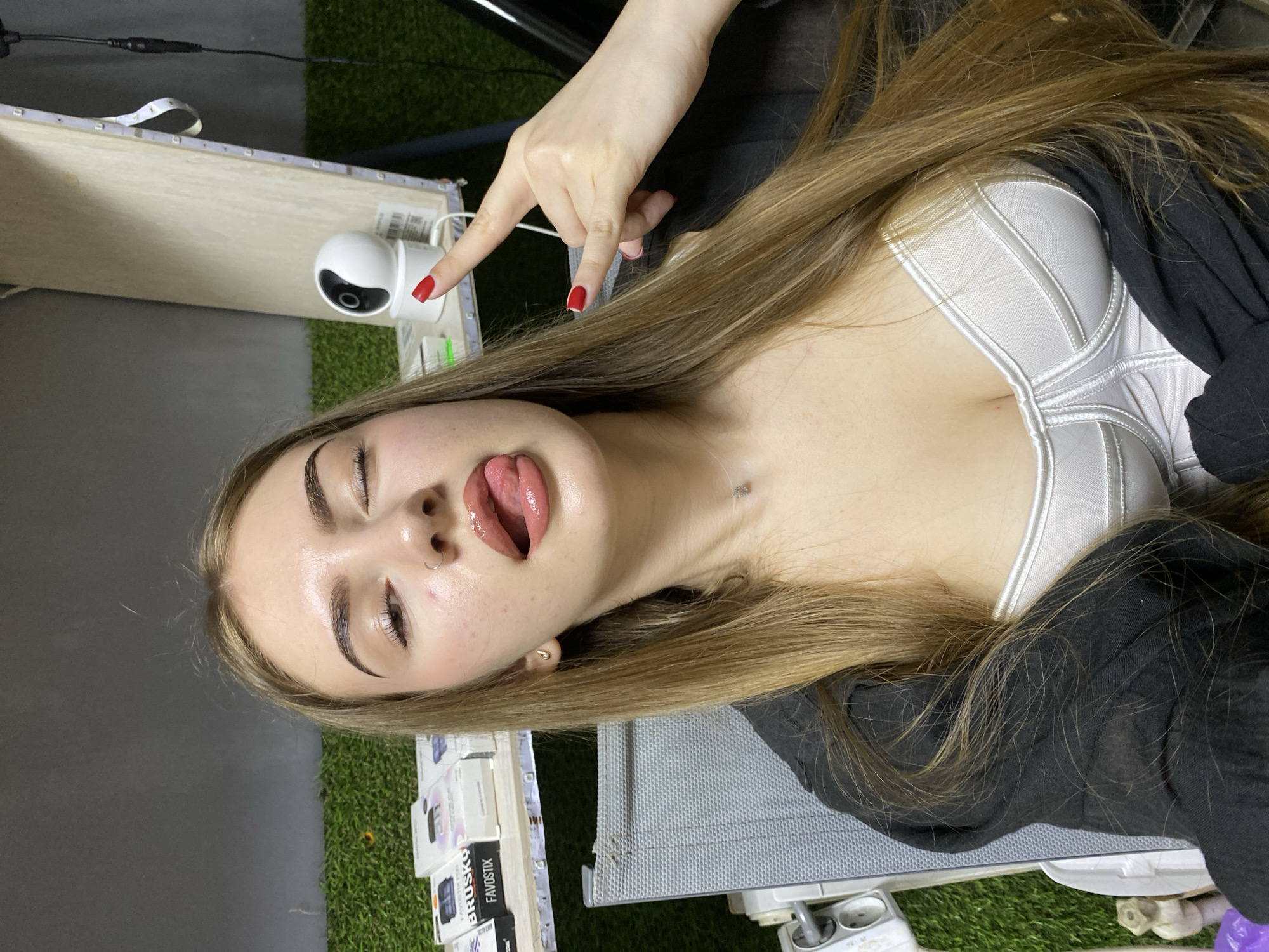 A young woman with long blonde hair and a white corset top is sitting in a chair, sticking her tongue out and making a rock-and-roll hand gesture. She is wearing makeup, including lip gloss and eyebrow pencil, and has a nose ring and earrings. The background appears to be an indoor setting with shelves containing various products, a security camera, and artificial grass. The lighting is bright, illuminating the woman's face and upper body. The specific location is not identifiable from the visual information provided in the image.