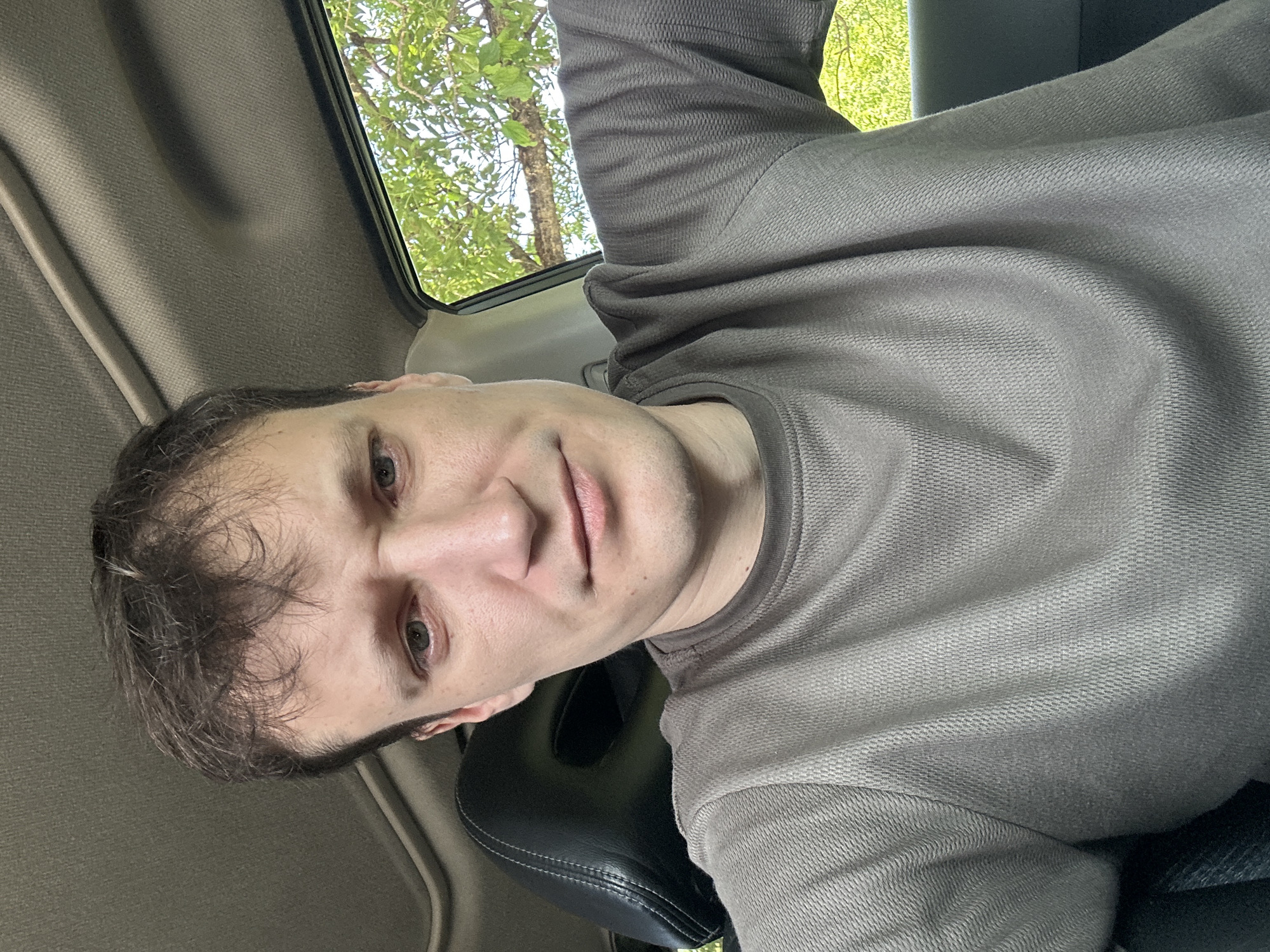 The image is a selfie of a man taken inside a vehicle. The man is looking directly at the camera with a neutral expression. He has light skin, brown hair, and green eyes. He is wearing a grey t-shirt. The interior of the car is visible, including the car's roof and a headrest. Outside the car window, green trees and foliage are visible. The lighting suggests it is daytime, and the setting appears to be outdoors, possibly in a rural area given the greenery. The exact location within Makiv, Ukraine, cannot be determined from the image alone.