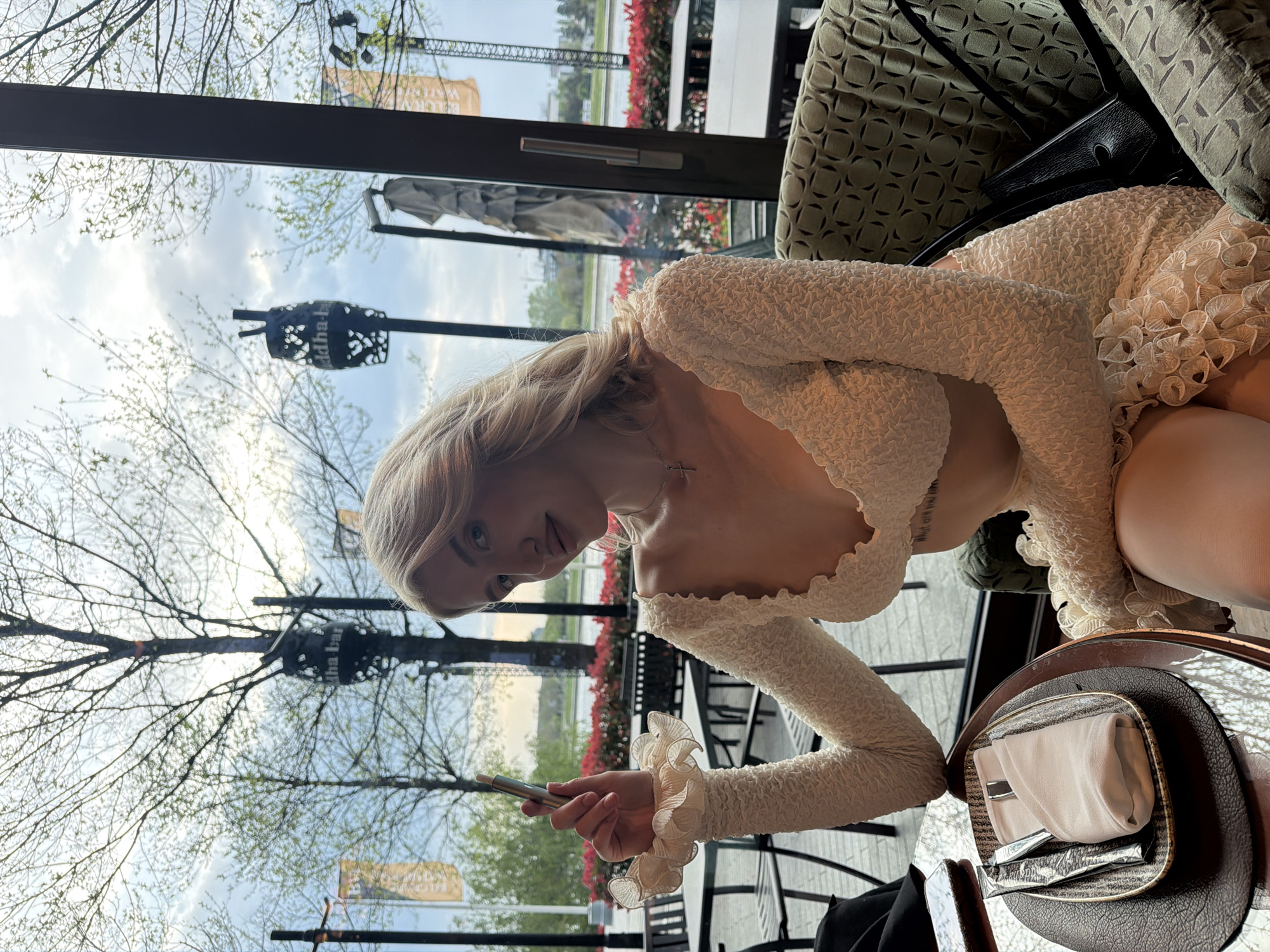 A fair-skinned woman with blonde hair is seated at a table in what appears to be an upscale restaurant in Belgrade, Serbia. She is wearing a white, long-sleeved dress with ruffled details at the neckline and cuffs. She is holding a silver-colored electronic cigarette in her right hand.
The table setting includes a patterned plate with a neatly folded white napkin, silverware, and sugar packets. Outside the window, trees, greenery, and what appears to be a racetrack are visible. A sign with the words "buddha-bar" can also be seen through the window, indicating the restaurant.
