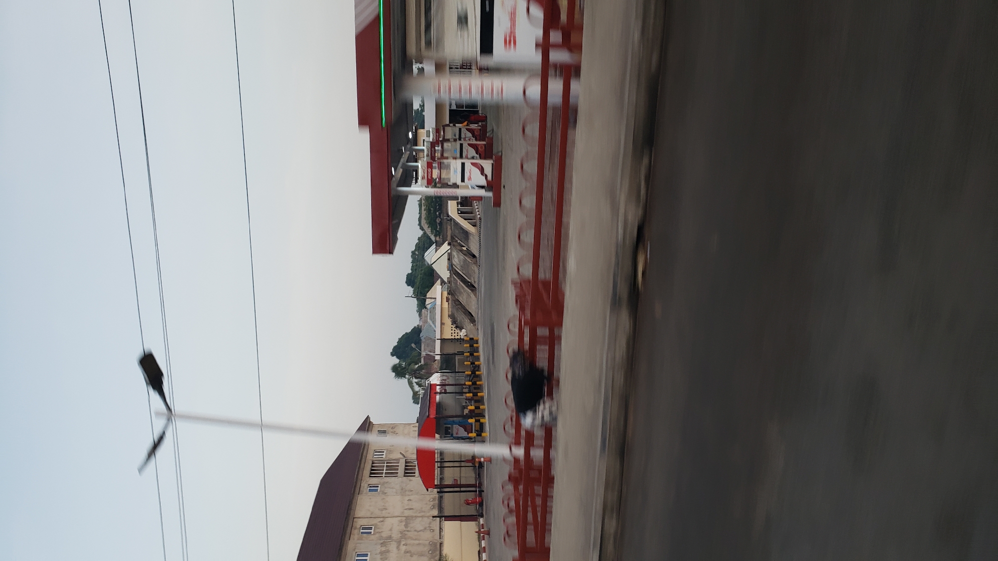 The image shows a gas station in Jalingo, Nigeria, from the perspective of a moving vehicle. The gas station features multiple pumps under a red canopy with green accent lighting. To the left, a light-colored building with a brown roof stands, and adjacent to it is a covered area with red and yellow bollards. A dark-colored object, possibly an animal, is visible on the red fence in the foreground. The road in front of the gas station is paved, and the sky is a pale blue with a few thin clouds. Power lines are visible stretching across the sky, supported by a lamppost on the left.