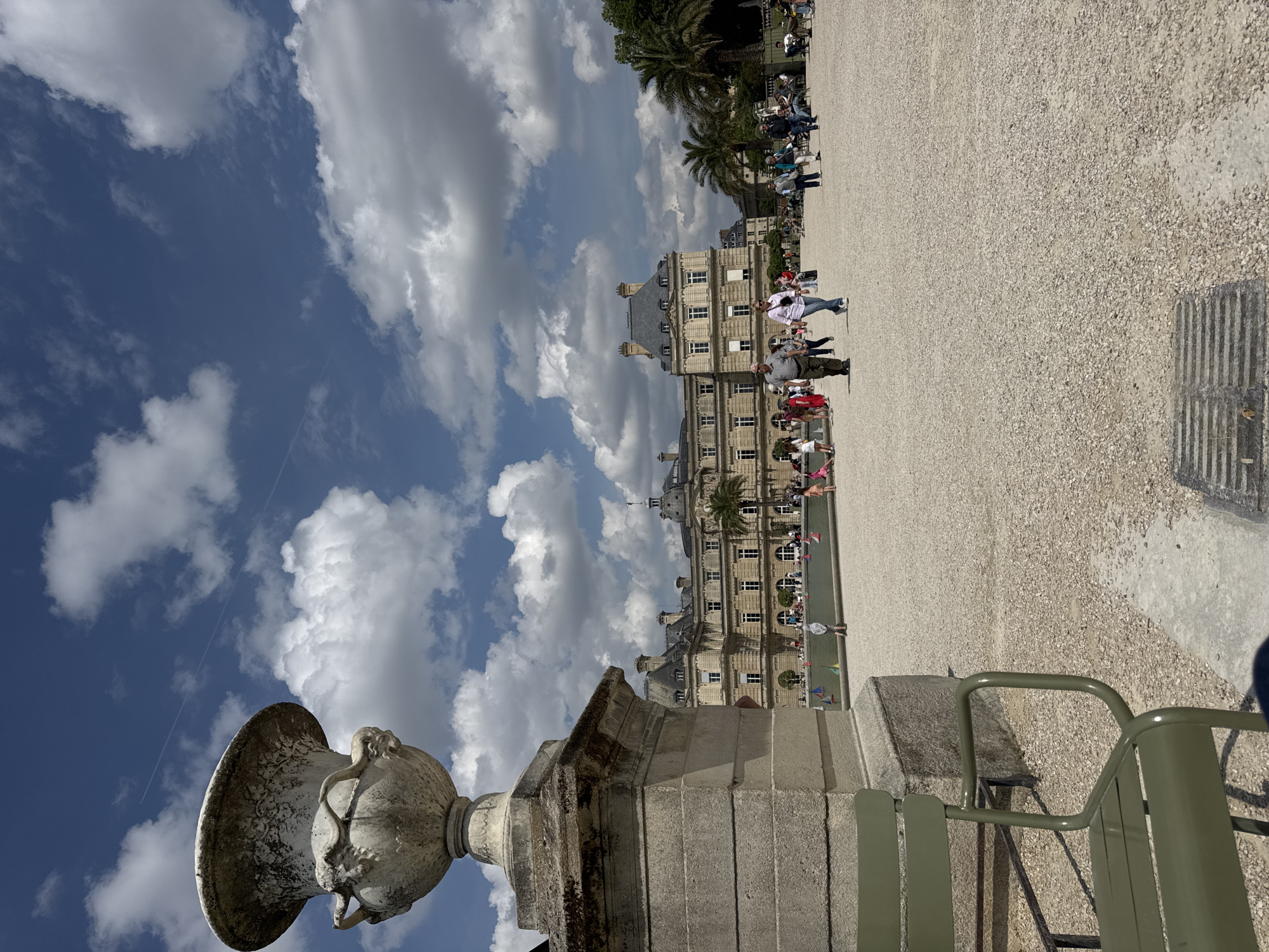 This image captures a scene in the Jardin du Luxembourg in Paris, France. The perspective is from a low angle, showcasing a stone vase on a pedestal in the foreground. The vase is intricately decorated and appears weathered. Next to the vase, part of a green metal park bench is visible. In the background is a view of the Luxembourg Palace, a grand building with multiple stories and a gray roof. Numerous people are scattered across the gravel pathways in front of the palace, suggesting a busy day. The sky is bright blue with scattered white clouds.