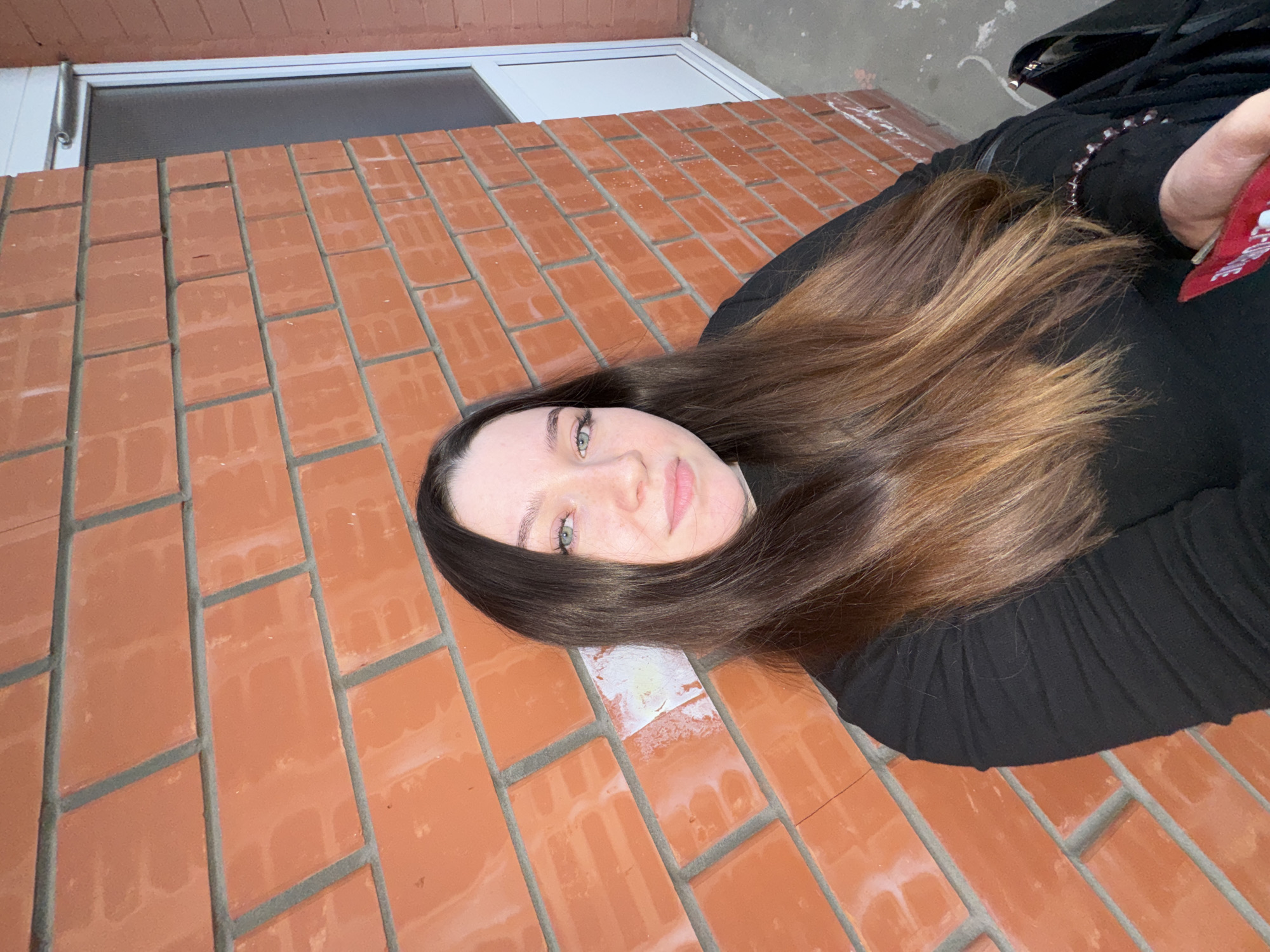 A young woman with long, brown hair and green eyes is posing for a selfie against a red brick wall. She is wearing a black long-sleeved shirt. To her right, a white framed door with a glass pane is visible. The ground beneath her appears to be concrete. The scene is outdoors, with natural lighting casting shadows. The presence of a red brick wall and the specific architectural style of the door and building could potentially place this scene in Perm, Russia, though no definitive landmarks are visible to confirm this.