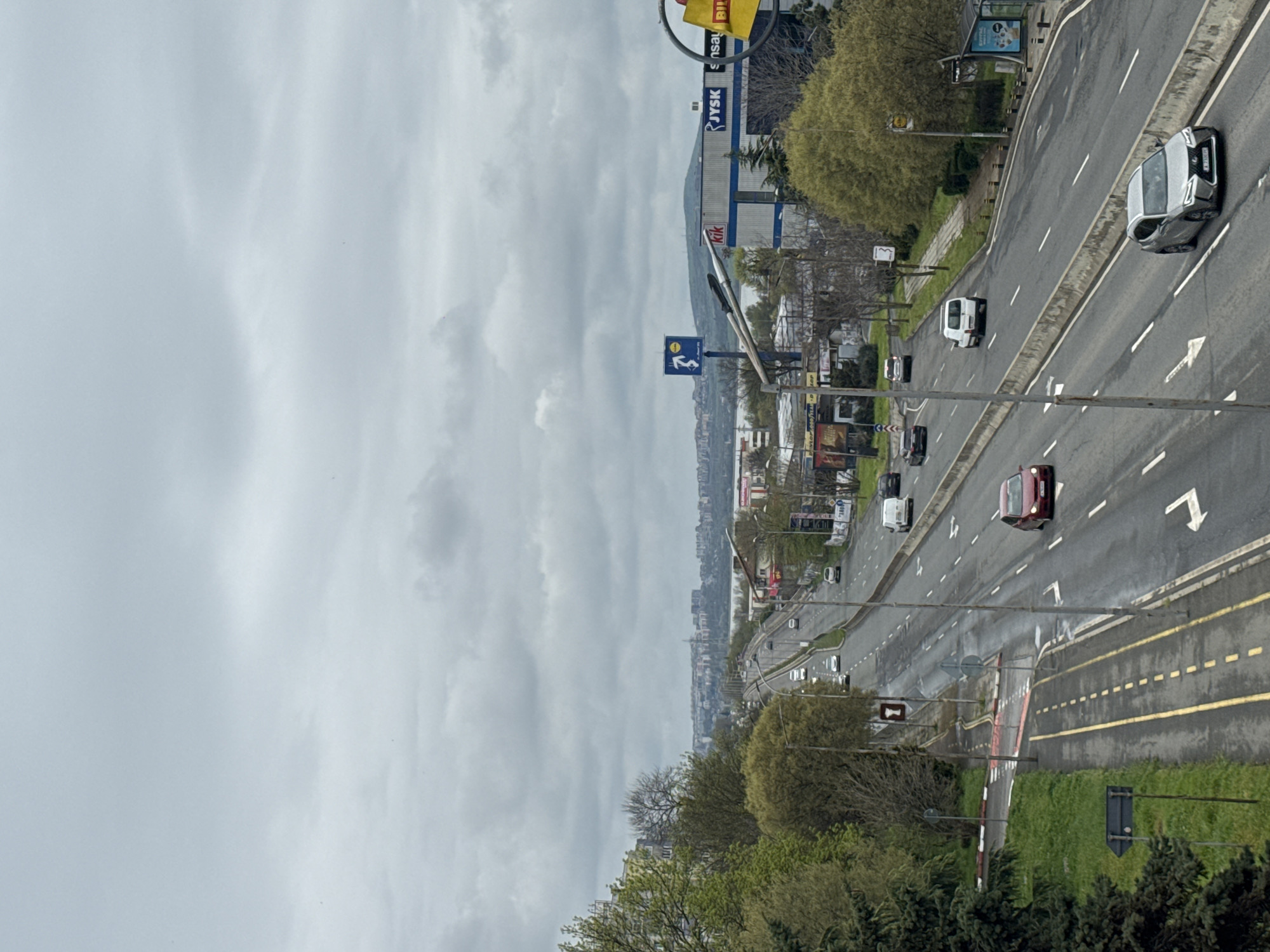 This image provides an elevated perspective of a multi-lane urban road in Burgas, Bulgaria, under an overcast sky. The road stretches into the distance, lined by trees and buildings, with a hazy cityscape or coastline visible on the horizon beneath the dense cloud cover.
Several vehicles are present on the road, including a white van, a silver car, and a red car, along with other vehicles further down the road, indicating active traffic. The road features clear white dashed lane markings, white directional arrows, and solid yellow lines defining the median and edges. Parts of the asphalt appear slightly wet or reflective, possibly due to recent rain.
Along the right side of the road, large commercial buildings are visible, displaying prominent signs. Notable visible text includes "JYSK," "KIK," and partially visible "Billa" and "Goodyear." A blue road sign with a white stylized figure is also present on the right. Green trees and grass are interspersed along the road, on the median, and in the foreground, providing natural elements within the urban setting. Streetlights are visible along the roadside. The scene depicts a typical busy daytime urban environment.