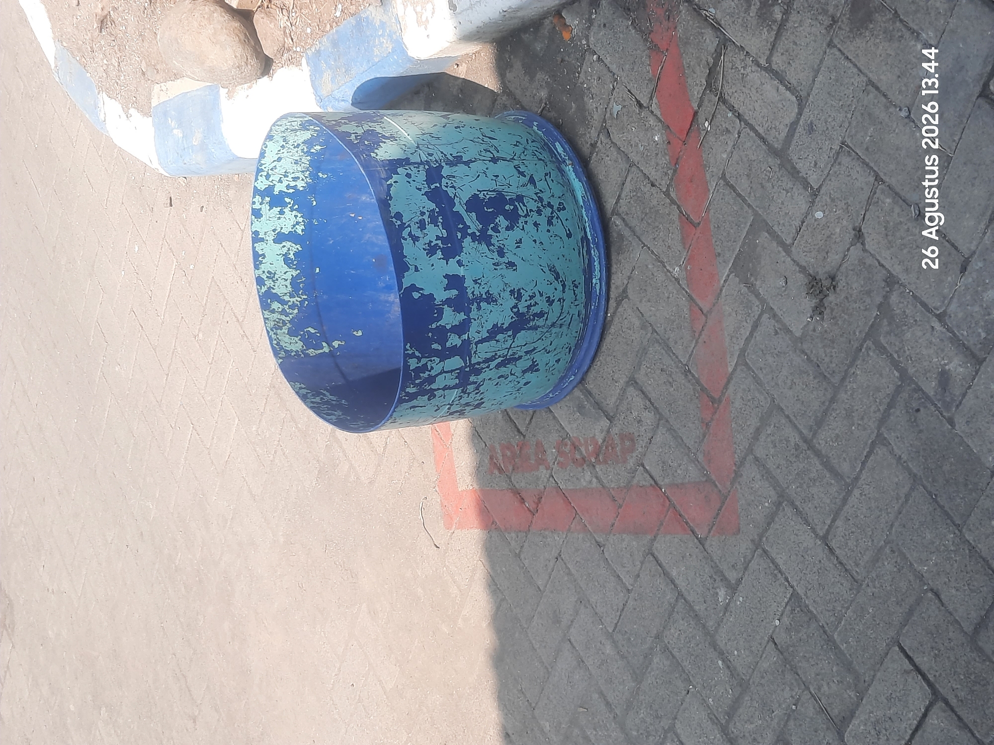 In Gending, Indonesia, a blue and teal cylindrical trash can with peeling paint sits on a herringbone-patterned brick paver surface. Red painted lines mark a designated area on the pavers, adjacent to a curb that is painted with alternating blue and white stripes. The scene is illuminated by natural daylight, casting a shadow from the trash can onto the brick surface. In the background, small stones and dirt are visible next to the curb. The bottom right corner of the image displays a timestamp: "26 Agustus 2026 13.44".