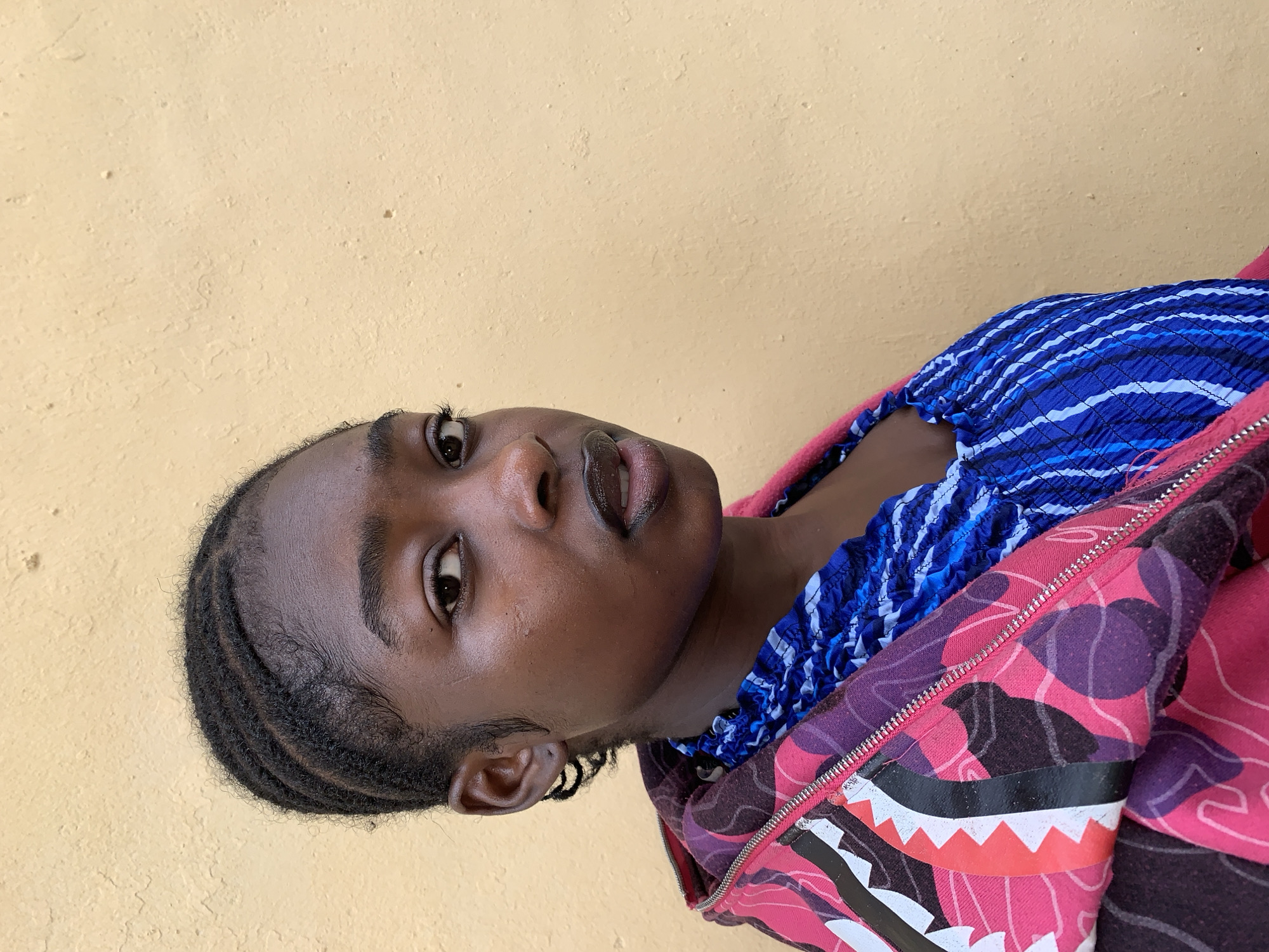 A young, dark-skinned individual with braided hair is looking up at the camera. The person is wearing a blue and white striped shirt and a pink and purple patterned jacket with a zipper. The background is a plain yellow wall. The picture appears to have been taken in Rafin Par, Nigeria.