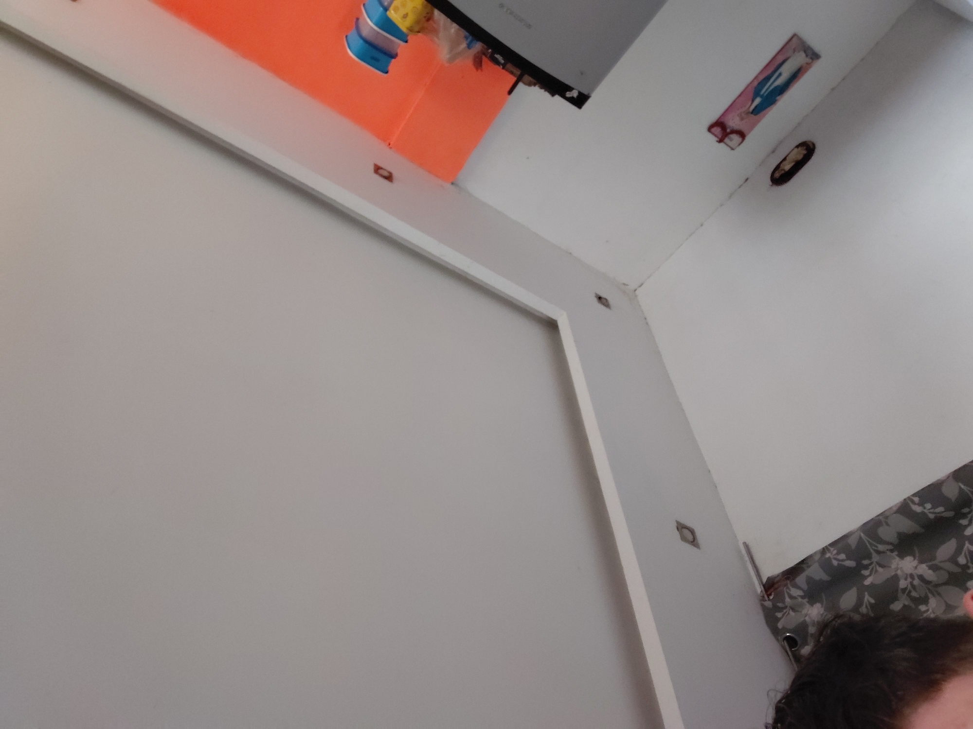 This image captures an indoor scene, likely a functional room such as a kitchen or utility area, viewed from a low, upward-looking angle. The perspective is tilted, showing white walls and a ceiling at an oblique angle.
In the upper central and left sections, a bright orange surface, possibly a shelf or the underside of a cabinet, is prominent. Resting on this orange surface are various items, including a blue and yellow plastic container, which might be a water bottle or a storage box, and several crumpled plastic bags. Above the orange surface, a grey-silver appliance, resembling a small refrigerator or microwave, is visible, seen from its underside. The brand name "GORENJE" is faintly legible on its surface, appearing inverted.
The left side of the frame is dominated by a large, plain white wall or the surface of a door, edged by white trim. Along this trim and the adjacent wall, several small, dark, square-shaped objects, possibly magnetic catches or mounting hardware, are visible.
The upper right portion of the image features a white ceiling and the corner where it meets the wall. A small, rectangular sign or sticker is attached to the ceiling, displaying a blue figure, possibly religious, and a partial letter "B" on its left side. Nearby, an oval-shaped, dark recess or hole is present in the wall or ceiling. Another small, dark, square object is affixed to the white wall in the lower right area.
The bottom right corner reveals a portion of a person's head, showing dark hair, suggesting the photo was taken by or includes the photographer. Next to the person's head, a patterned curtain, grey with a white floral or leaf design, is visible.
The scene depicts an everyday domestic or utility environment. The image was captured in Santa Ana, Venezuela.