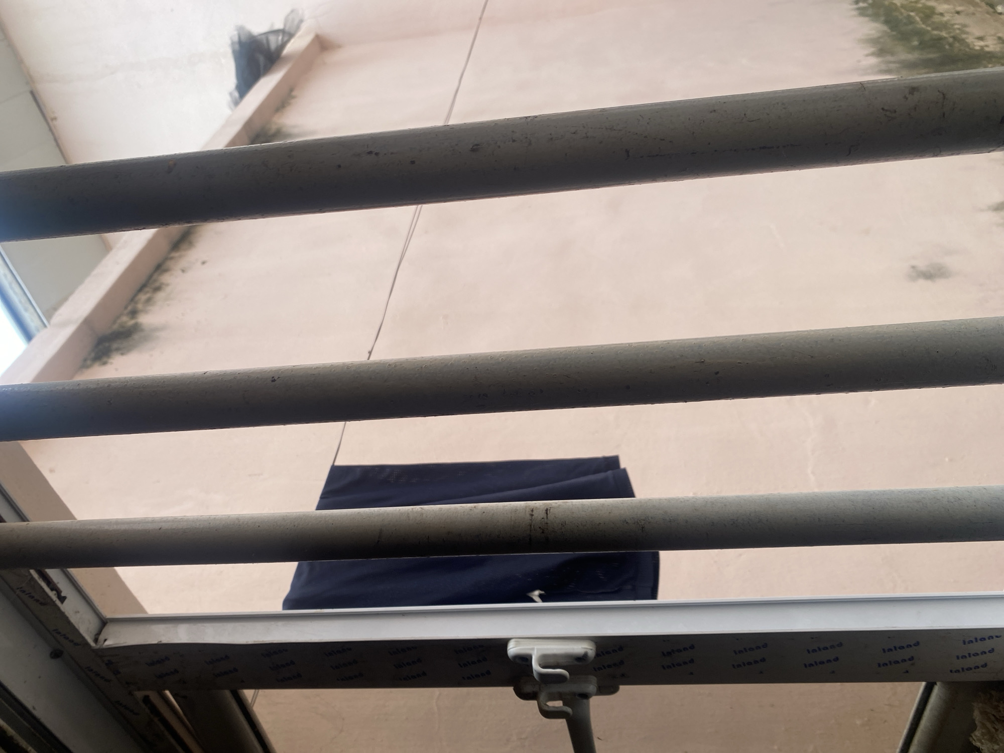 The image shows a view looking up towards a pale pink wall, likely an exterior building wall, from a lower vantage point. Three horizontal grey metal pipes or bars are prominently positioned across the frame, creating a layered effect. Between the second and third pipe from the top, a dark blue piece of fabric, possibly clothing, is hanging. The wall appears weathered, with patches of dark discoloration, suggesting it is outdoors and has been exposed to the elements. The top of the frame shows a sliver of a light-colored ceiling and a dark, indistinct object in the upper left corner, possibly a net or screen. The lower part of the image reveals a white window frame with repeating text that is partially obscured and difficult to read clearly, but some fragments appear to say "Jaland," "Taland," and "Inland." The overall impression is a somewhat neglected outdoor setting, perhaps a balcony or a backyard, with laundry hanging to dry. The lighting suggests it is daytime.