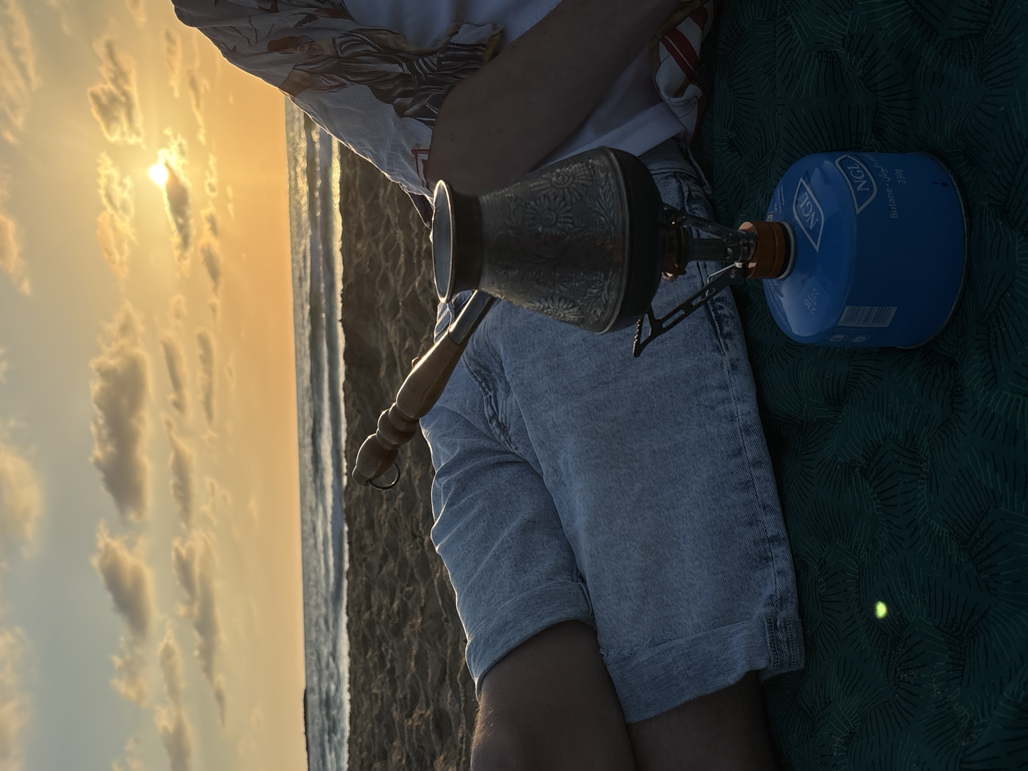 A person is sitting on a patterned dark green mat on a beach. They are wearing a white and patterned shirt and light-blue denim shorts. They are using a portable camping stove with a blue gas canister to heat a metal cezve with a wooden handle. The sun is setting over the ocean in the background, creating a warm golden light. The location is likely near Hof HaCarmel Regional Council, Israel, due to the coastal environment.