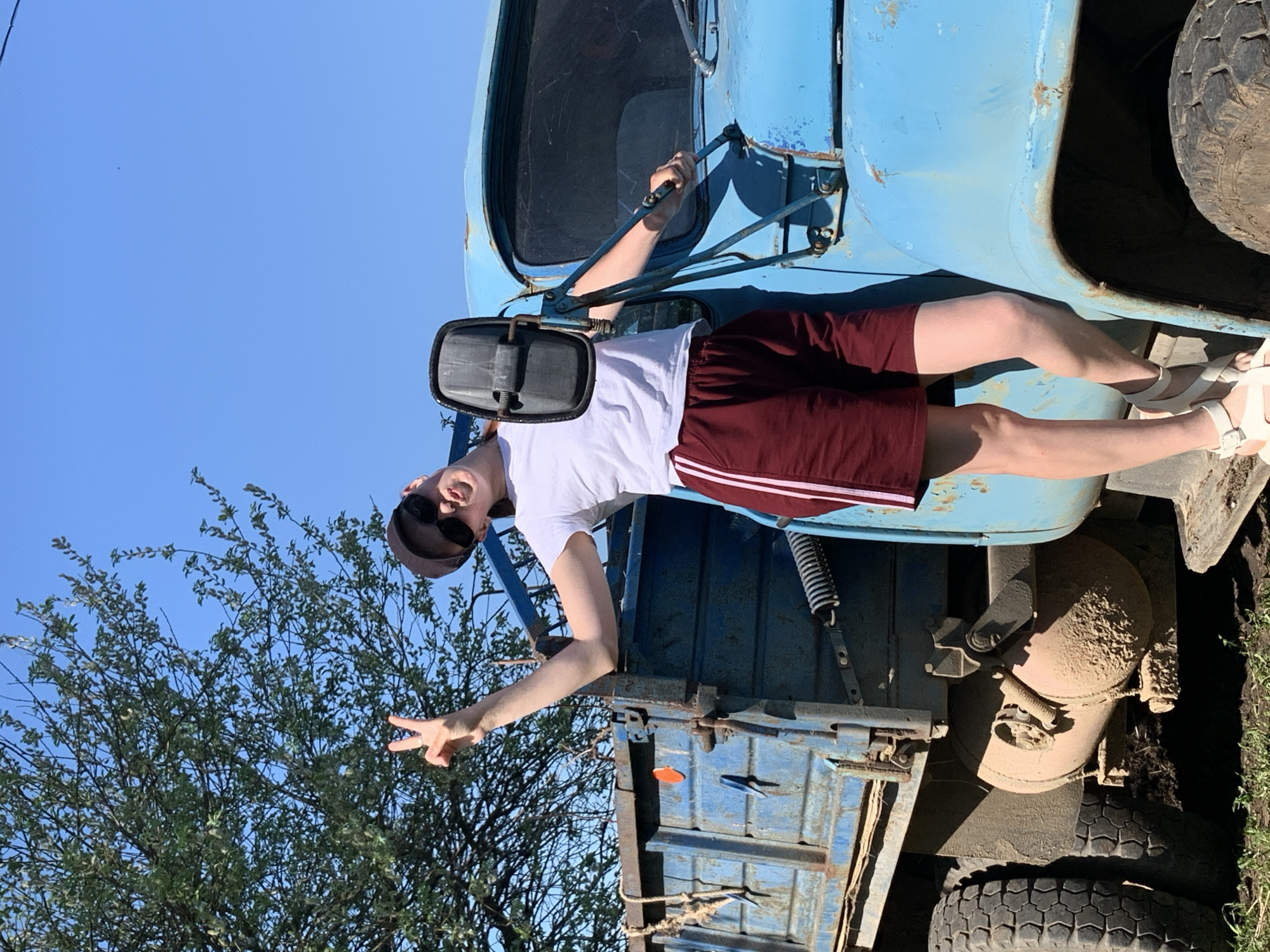 A person is standing on the side of a light blue truck with a dirty chassis and tires. The person is wearing a white shirt, maroon shorts with white stripes, and sandals. They are also wearing a cap and sunglasses, and holding onto the truck's side mirror. They are making a peace sign with one hand. To the left of the truck, there is a tree with green leaves. The sky is blue. Based on visual information, the location is not identifiable as Фоки, Russia.