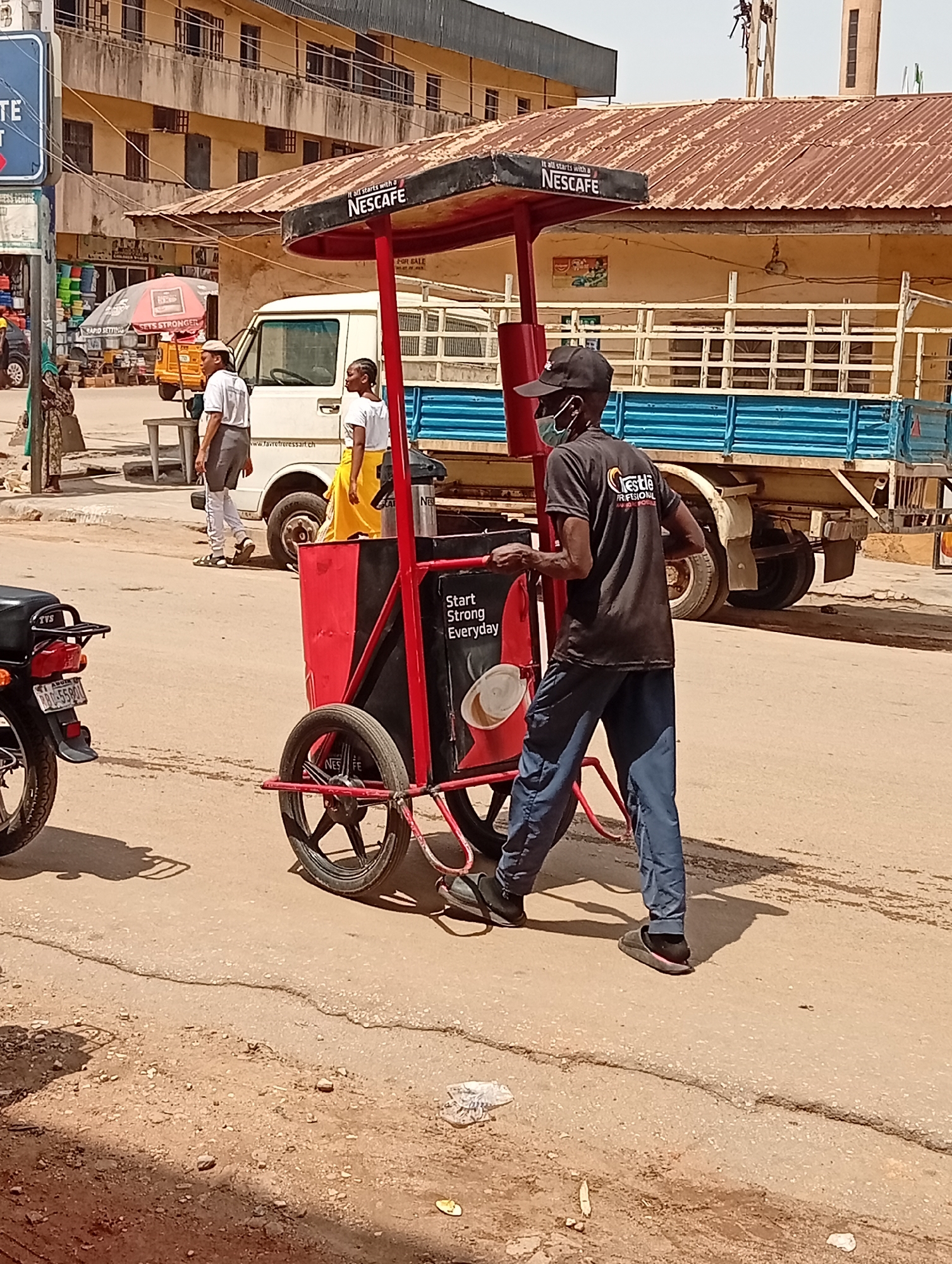 A man wearing a black baseball cap, a black t-shirt with a Nestle logo, blue pants, and black sandals is pushing a red and black mobile Nescafe cart on a paved street in Maiburiji, Nigeria. He is also wearing a face mask. The cart has a sign that reads "Start Strong Everyday." A white van and a blue flatbed truck are parked in the background. Two people are walking near the van. A black motorcycle is parked on the left side of the frame. Buildings with rusted roofs and windows are visible in the background, suggesting an urban setting. A street sign is visible in the top left corner, and a Nescafe advertisement is prominently displayed on the cart's canopy. The street surface is cracked and shows signs of wear.