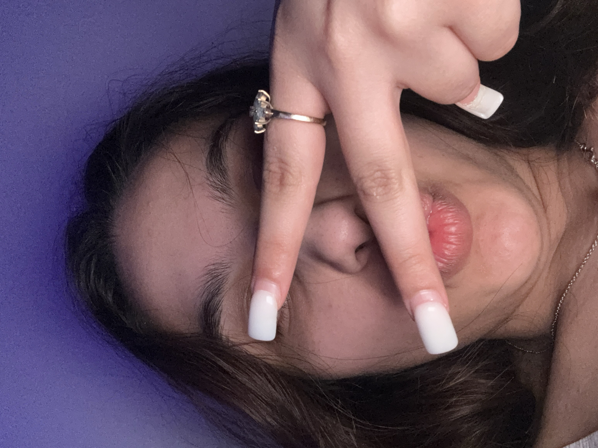 A close-up, angled shot depicts a young person's face obscured partially by a hand displaying long, square-shaped, white acrylic nails. A ring adorns the ring finger, featuring a light-colored gemstone surrounded by smaller stones on a gold band. The person has brown hair and visible eyebrows, with closed eyes. The nose and lips are centrally positioned in the frame, exhibiting a light pink hue. A silver chain necklace is partially visible on the right side of the neck. The background appears to be a plain, solid purple color. The image location and city are not discernible from the visual information provided.