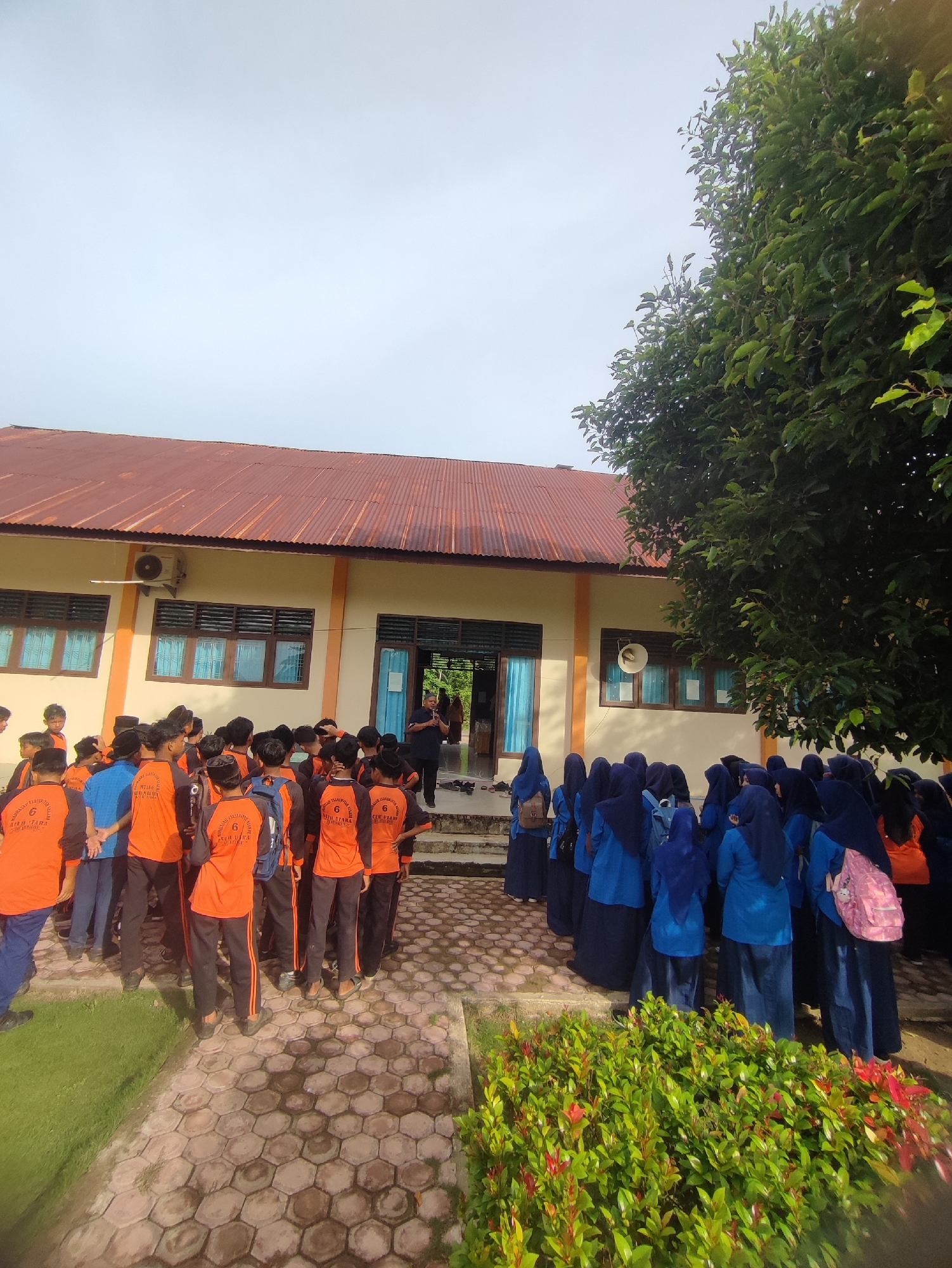 In Blangriek, Indonesia, a group of students, predominantly wearing orange shirts with black sleeves, stand gathered outside a light-colored building with a rust-colored corrugated metal roof. The building has several windows with dark blinds. To the right of the building, a large tree with dense green foliage partially obscures the sky, which is overcast.
The students are arranged in a loose cluster, facing the building and a central figure who appears to be an adult, possibly a teacher or instructor, dressed in a dark shirt and dark pants. Some students are looking at the adult, while others are interacting amongst themselves or looking in different directions. A few students are wearing blue headscarves, and one has a pink backpack.
The ground in front of the building is paved with hexagonal concrete tiles, and to the left, there is a patch of green grass. In the foreground, a low-lying bush with bright red and green leaves is visible. An air conditioning unit is mounted on the side of the building, and a speaker is visible above one of the windows. The scene appears to be taking place during the daytime, under natural light.