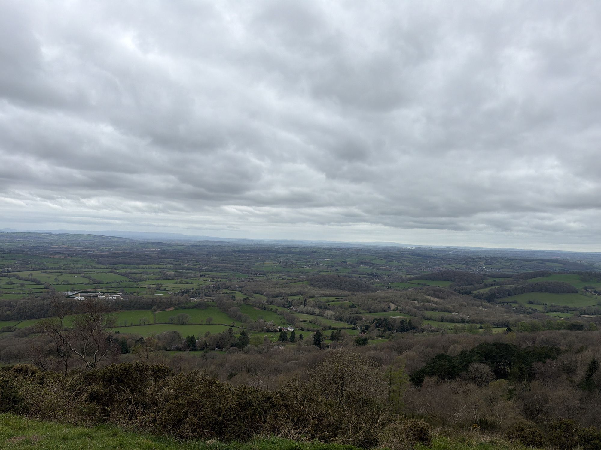 The image presents a vast landscape from an elevated vantage point in the Malvern Hills, United Kingdom. The scene is dominated by a cloudy, overcast sky. Below, a sprawling patchwork of green fields and wooded areas stretches into the distance. The landscape is punctuated by clusters of buildings, possibly farmhouses or small villages. The foreground shows vegetation, including scrubby bushes and grass, indicating the viewpoint's elevated position. There are no people visible in the frame. The overall impression is one of a rural, hilly landscape under a heavy, cloudy sky.