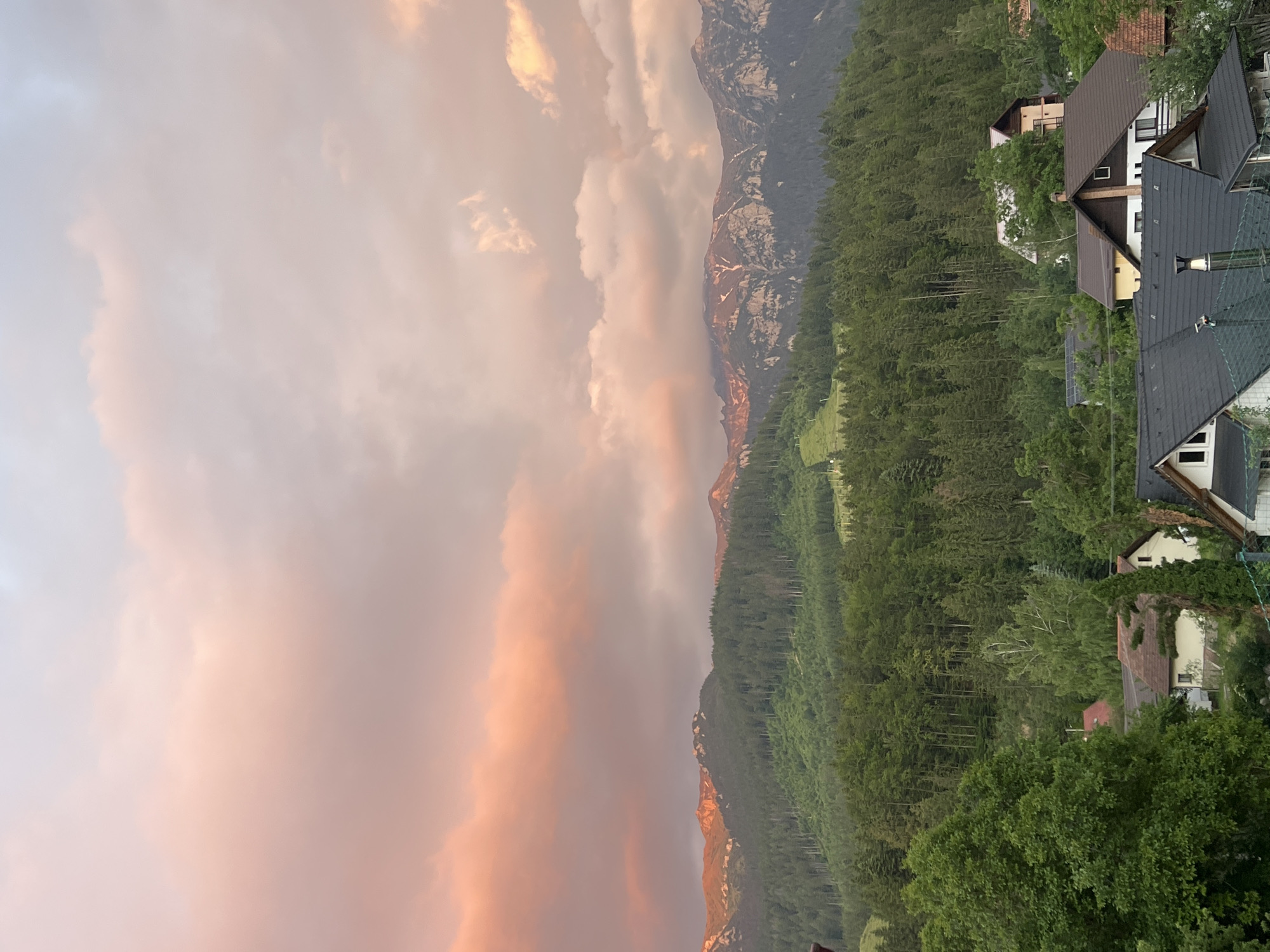 The image depicts a mountainous landscape at sunset, with houses nestled in the foreground. The sky is filled with soft, diffused light, casting a warm pink and orange glow on the clouds. The mountains are covered in dense green forests, with some rocky outcrops visible at their peaks. In the foreground, several houses with tiled roofs are scattered among the trees. The overall impression is one of tranquility and natural beauty. The scene is devoid of any people or animals.