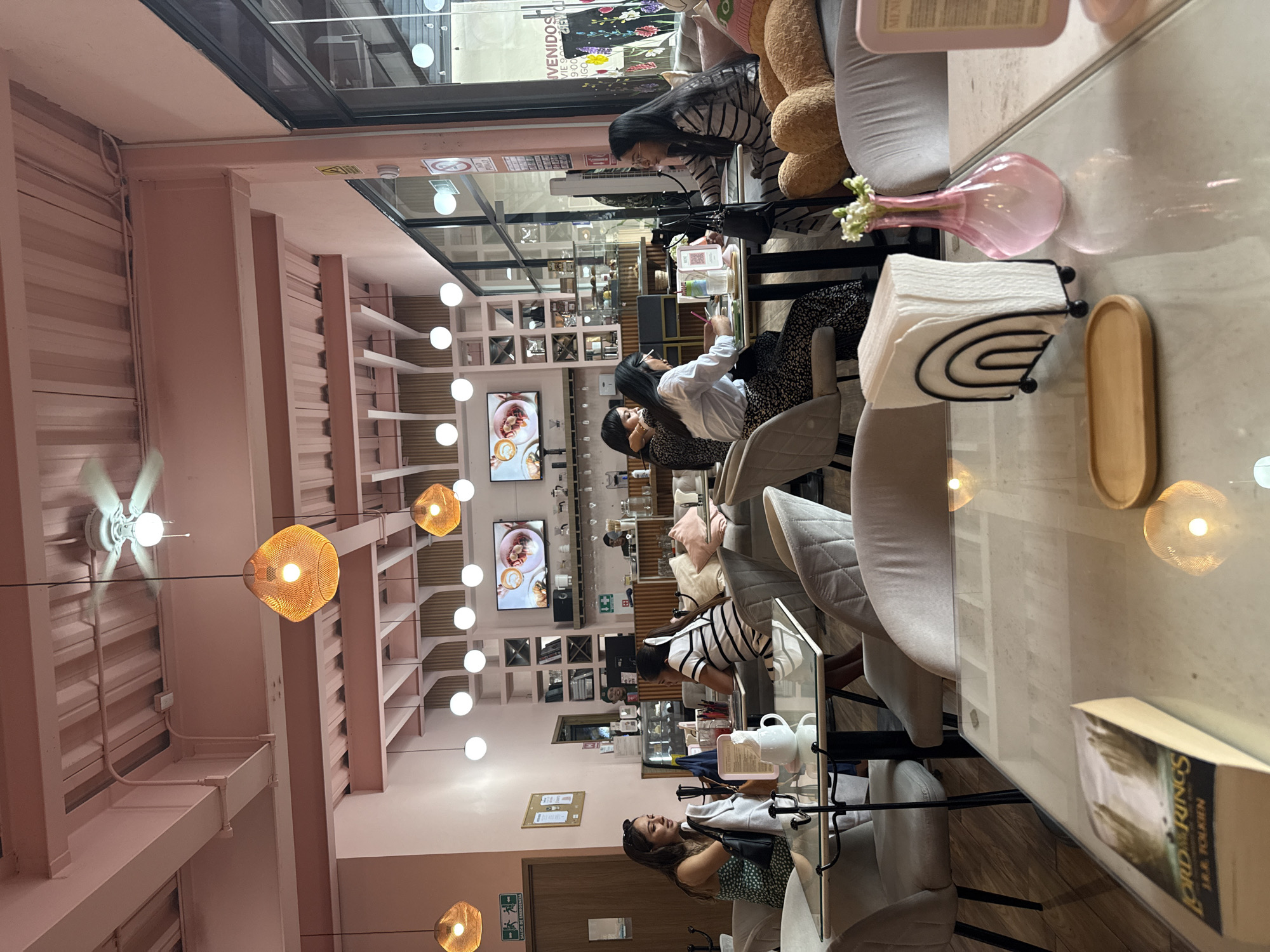 The image shows the interior of a cafe in Puebla, Mexico. The walls and ceiling are painted in pastel pink. There are light fixtures hanging from the ceiling. In the background, there's shelving and televisions displaying images of food. Several people are seated at tables along the length of the room. A book titled "The Lord of the Rings" is on the table. There are napkins, a vase with flowers, and dishes on the table.