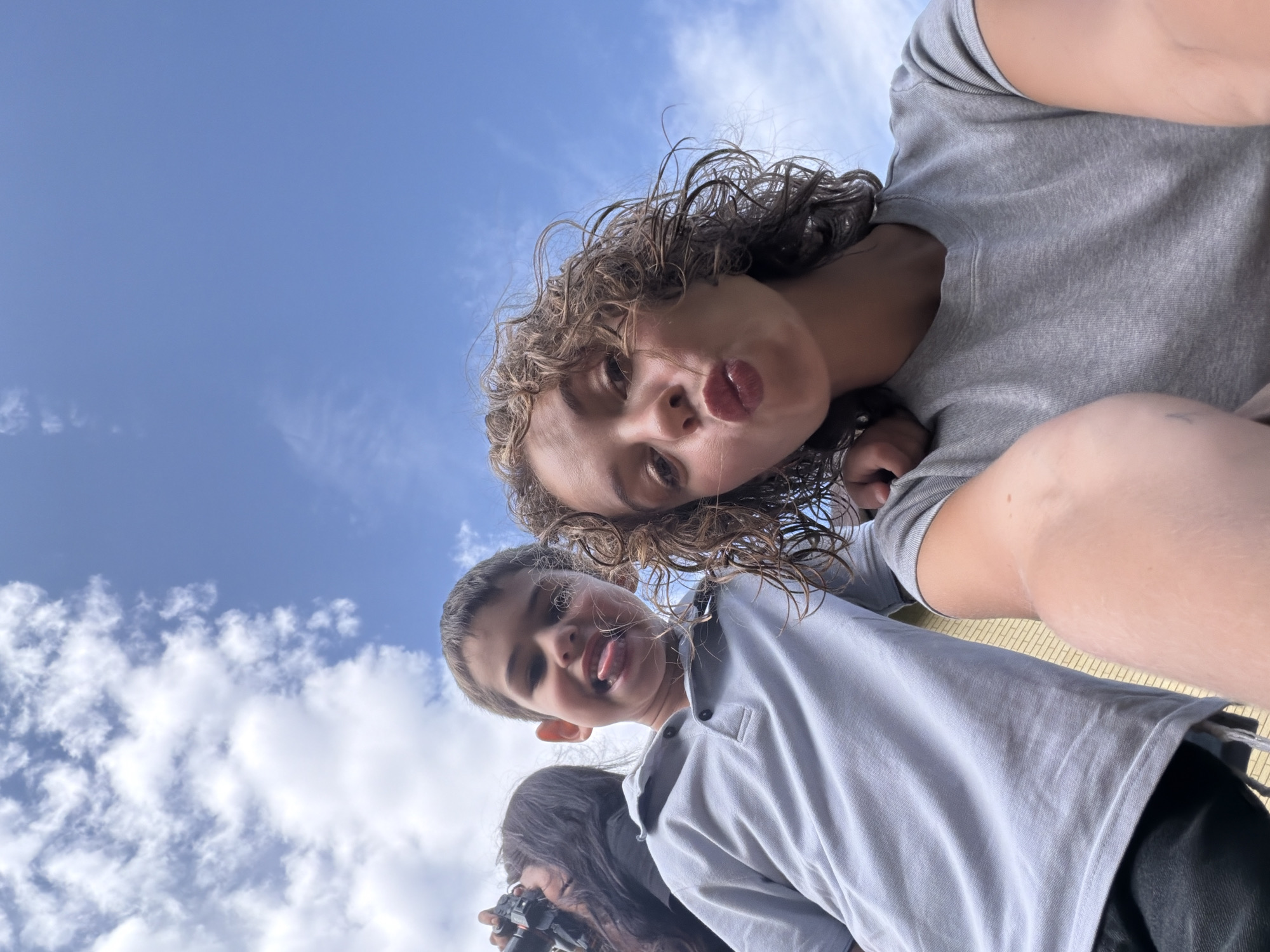A low-angle selfie taken outdoors against a blue sky with scattered white clouds. Two young people are the primary subjects. On the left, a boy with short brown hair wears a light gray polo shirt and sticks his tongue out. To his right, a girl with curly brown hair is puckering her lips and wearing a gray t-shirt. In the background on the far left, partially visible, is a person with dark hair holding a camera. The lighting suggests it is daytime. There is no indication of the specific location within Mishutkino, Russia.