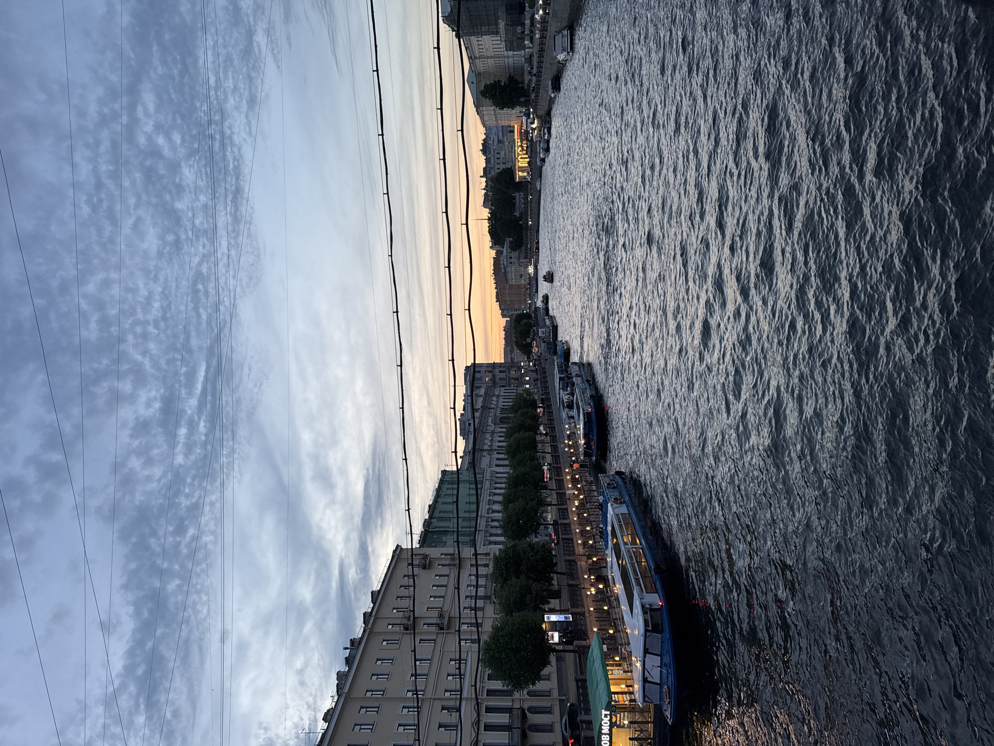 The image captures a wide shot of a canal in Saint Petersburg, Russia, during twilight. Several boats are visible on the water, including tour boats and smaller vessels. The canal banks are lined with buildings, some of which are illuminated, and trees. The sky is cloudy with a hint of orange from the setting sun. Utility wires are strung across the upper portion of the frame. The water is choppy with visible ripples and reflections.