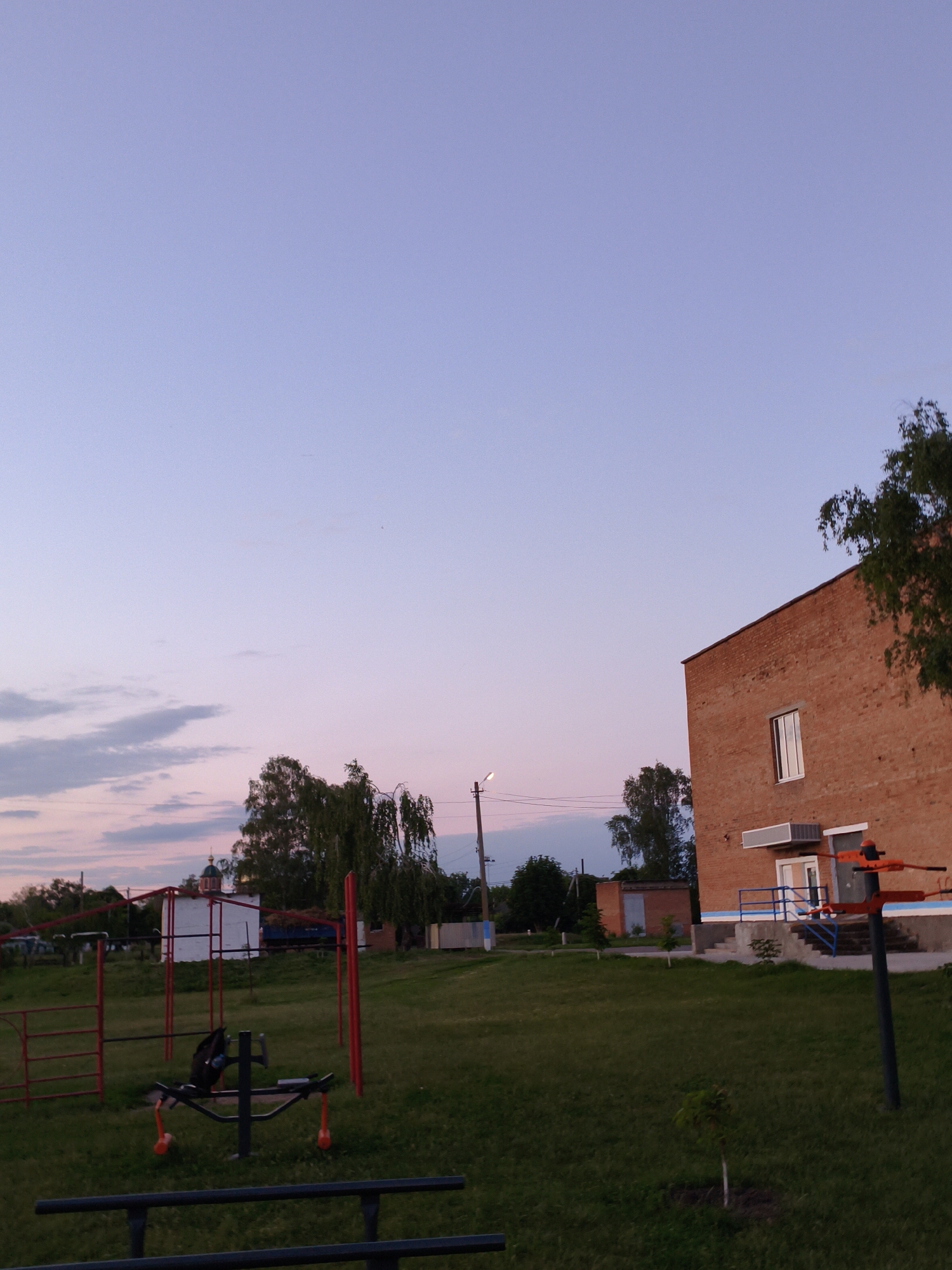 The image depicts an outdoor scene at dusk in the city of Ustyvytsia, Ukraine. In the foreground, there is a children's playground with a red frame and black metal bars. To the right, a brick building with a white window and blue trim is visible. A tree with green foliage is situated in front of the building. The sky is a gradient of light blue and pink, with scattered clouds. In the background, there are more trees and a small white building with a domed roof, possibly a church. The overall impression is one of a rural or suburban environment during twilight.