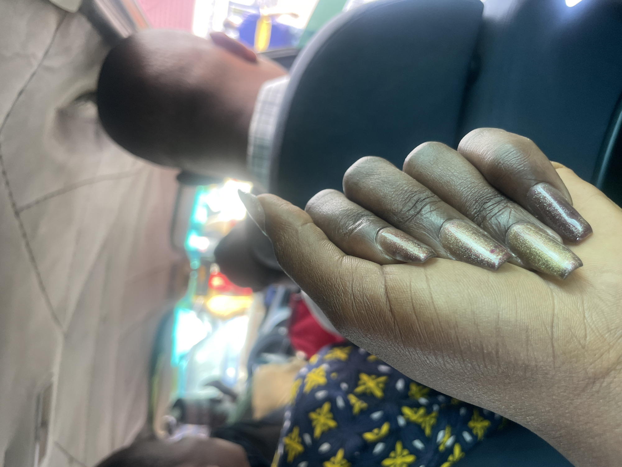 An over-the-shoulder shot focuses on a person's hand with long, almond-shaped fingernails painted with a shimmery brown polish. The hand is positioned in the foreground. In the background, blurred figures are visible inside a vehicle. One person in the middle ground, seen from the back, wears a dark jacket over a plaid shirt. Another person in the further background wears a blue shirt with yellow stripes. The interior of the vehicle, including the ceiling, is visible. The scene appears to be inside a vehicle in the city of Wauro Jabbe, Nigeria.