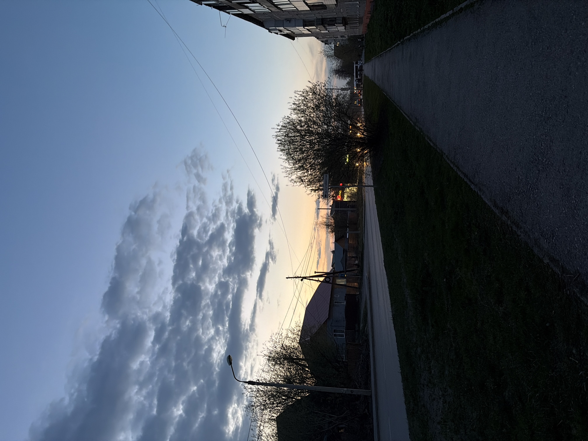 The image depicts a street scene in Perm, Russia at dusk. A paved sidewalk runs along the right side, bordered by grass. A multistory apartment building stands at the top edge of the frame. Overhead power lines stretch across the sky. In the distance, houses with red roofs are visible. Clouds fill the sky, partially obscuring the sunset. A street lamp is on the left. A tree with bare branches is in the middle ground.
