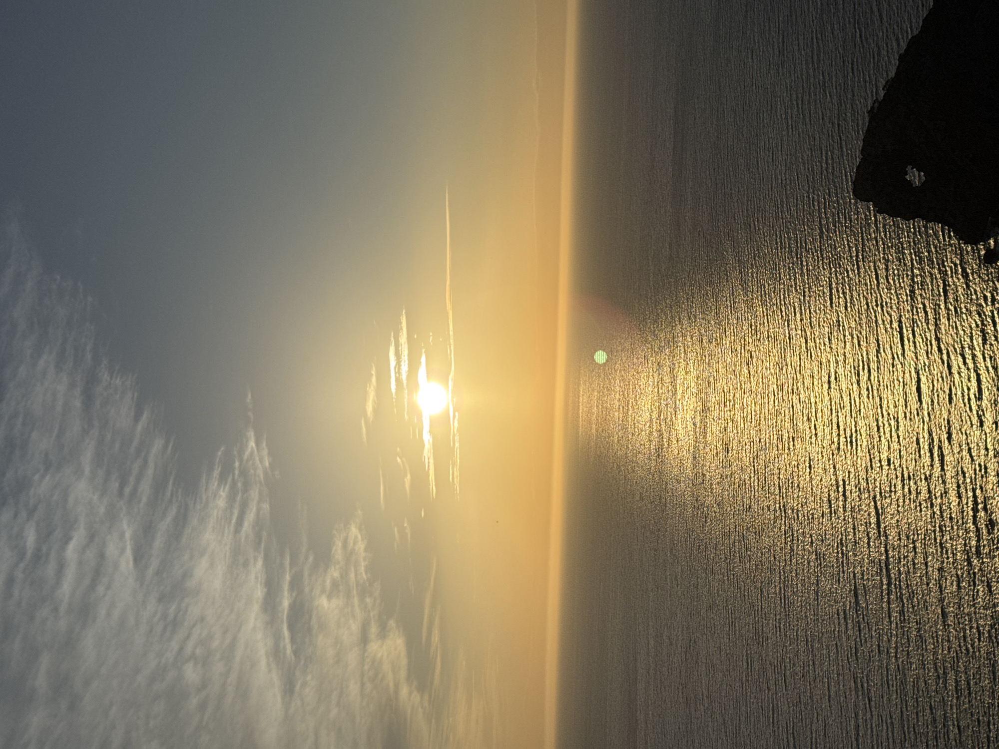 A photograph of the ocean horizon during sunset in Deià, Spain. The water is glistening gold from the sun. A rock is visible on the top right. Some clouds are to the left, and the sky is a grey-blue color. A slight lens flare of the sun is visible on the water.