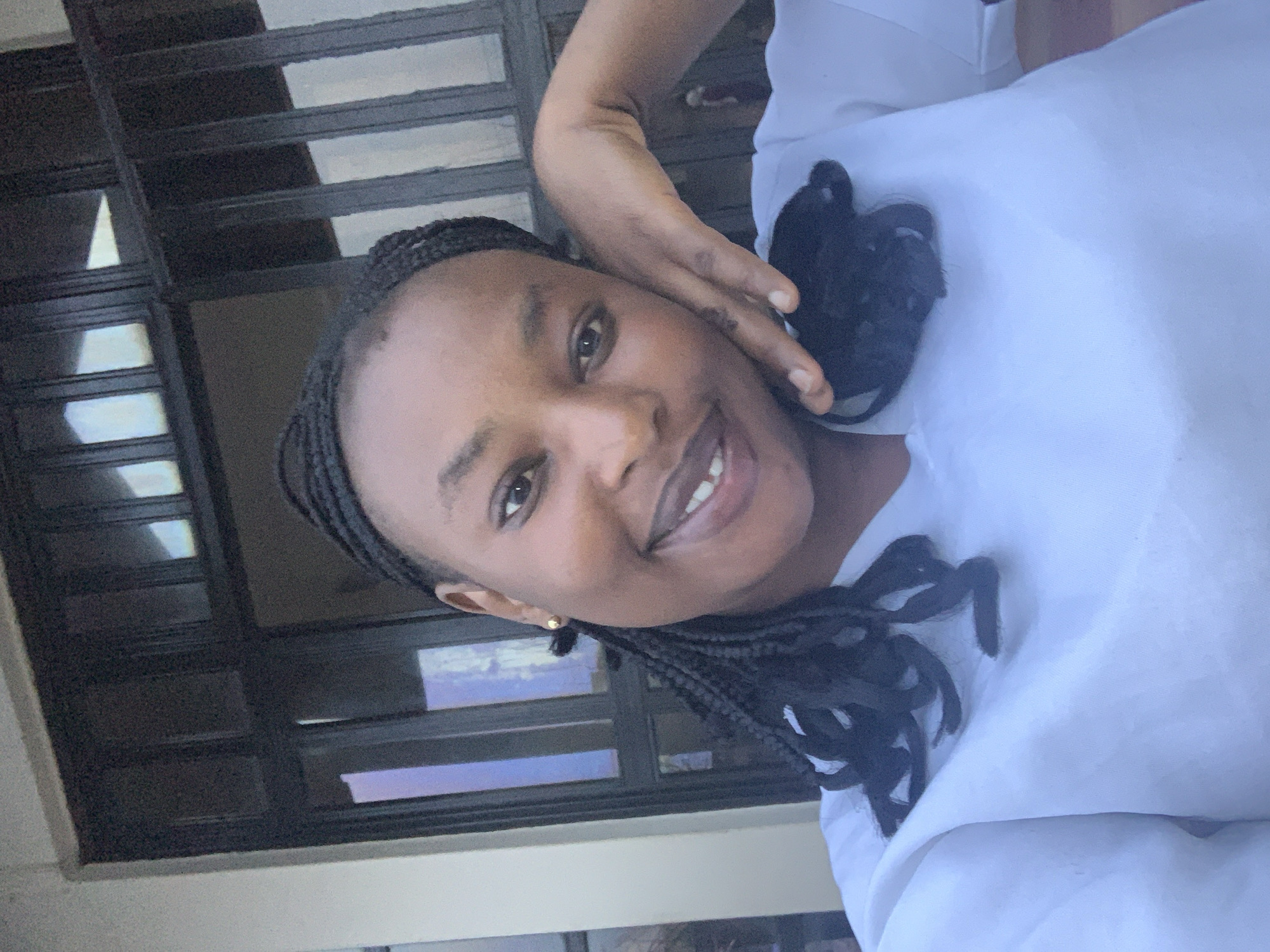 In Jos, Nigeria, a young woman with braided hair and a gold earring smiles at the camera, her right hand resting on her cheek. She is wearing a white top, and her hair is styled in black braids. Behind her, a dark-colored door with glass panes is visible, suggesting an indoor or semi-outdoor setting in Jos, Nigeria. The overall scene is a close-up portrait.