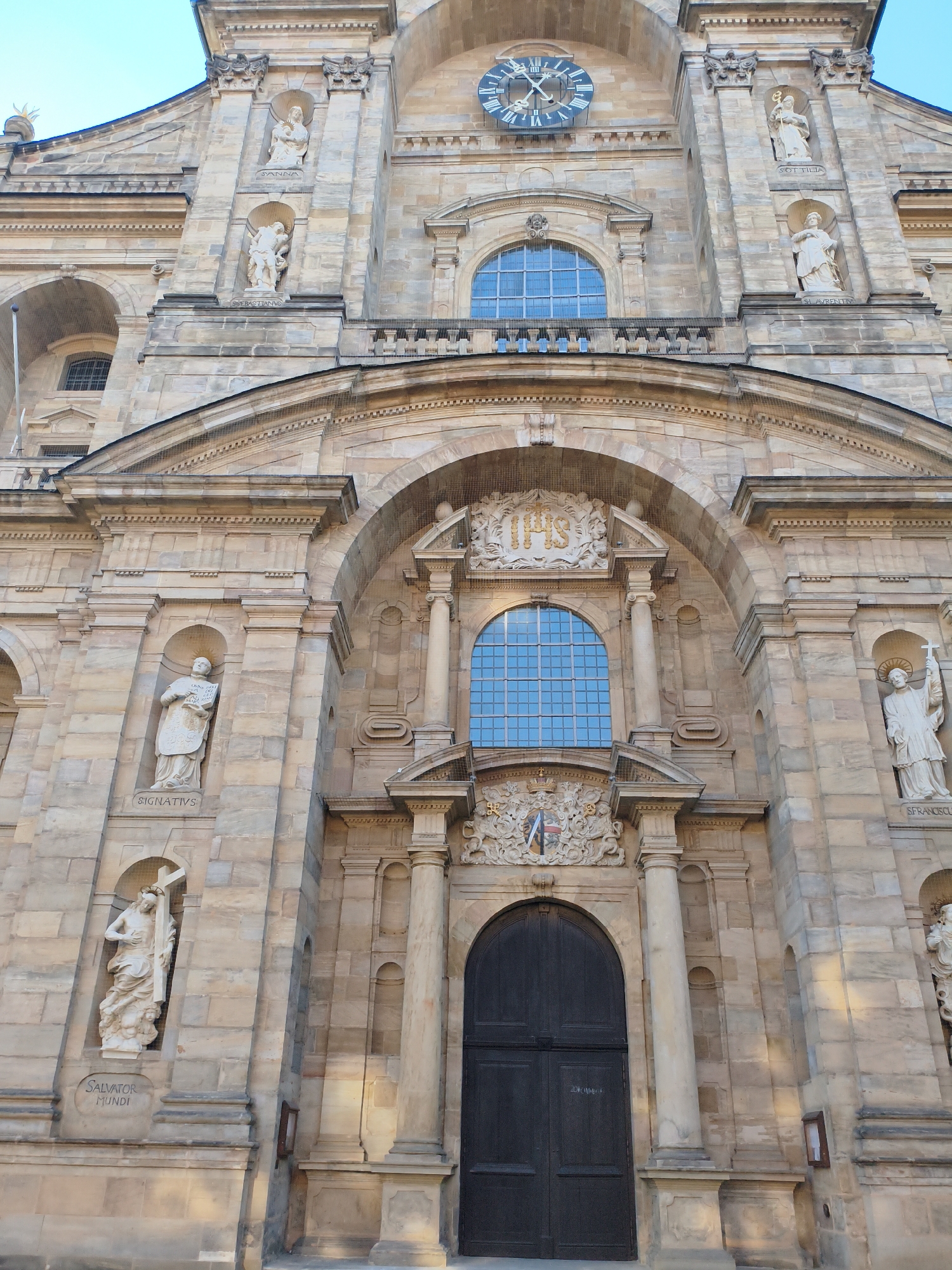 The image is a low-angle exterior shot of the facade of Bamberg Cathedral, located in Bamberg, Germany. The cathedral is constructed of light-colored stone and features elaborate architectural details. Prominently displayed on the facade are multiple statues of religious figures in niches, along with a large clock face above a semicircular window. The entrance to the cathedral is a large, dark wooden door beneath an arched entryway, flanked by columns. The sky is clear and blue. The photograph captures the grandeur and historical significance of the cathedral.