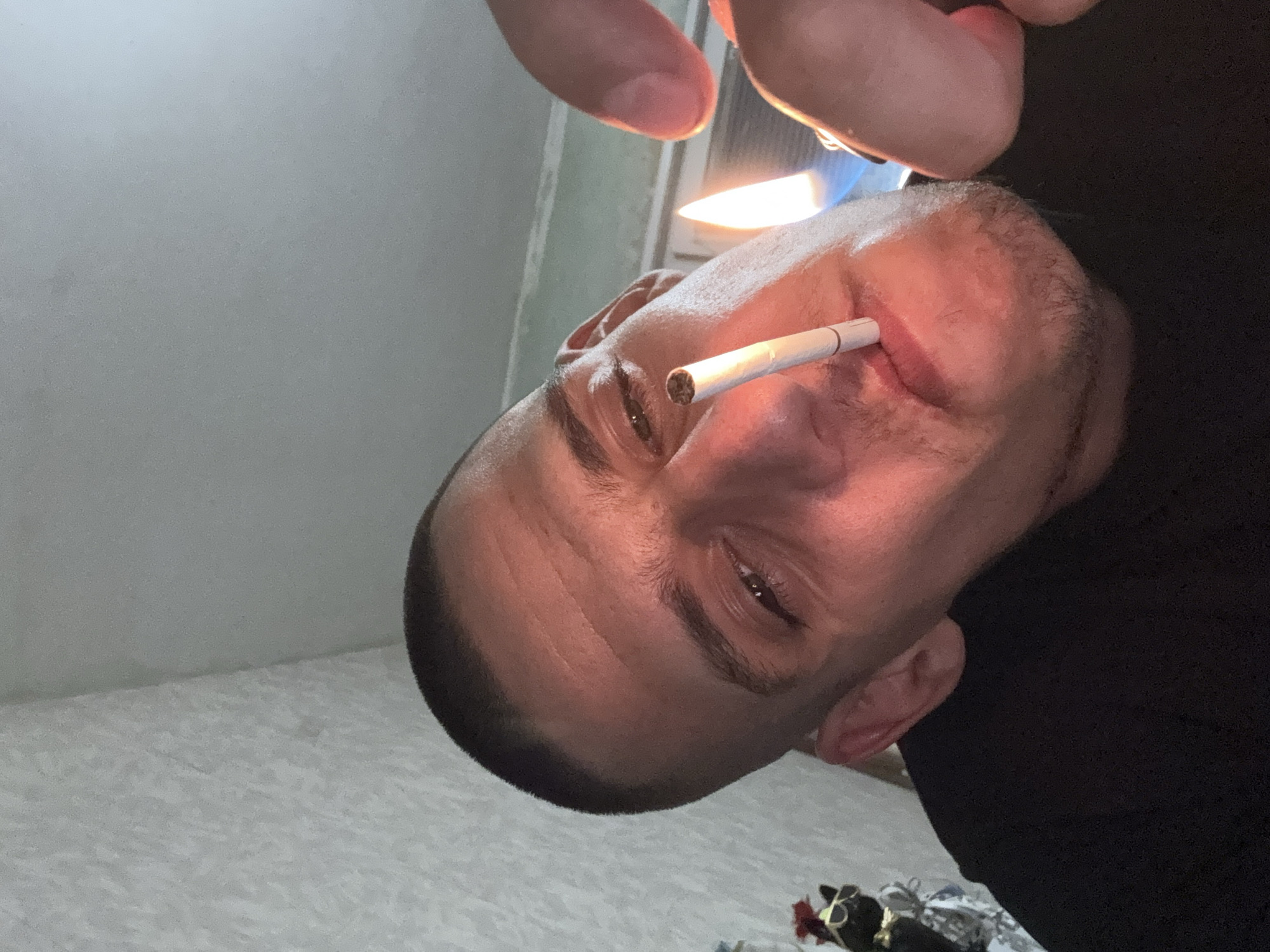 The image shows a close-up, downward-angle shot of a man lighting a cigarette. He has a shaved head and appears to be in his 30s or 40s. He is wearing a black t-shirt. In the foreground, a cigarette is held between his lips, and a lighter with a blue flame is being used to ignite it. The background is a wall with textured wallpaper. A small, dark figurine wearing sunglasses and a red bow can be seen on the left side of the image, partially obscured. The lighting is warm and casts shadows on the man's face, highlighting his features. The overall impression is that of a candid, personal moment captured indoors. The provided information does not specify a location within the city of Sofia, Bulgaria.
