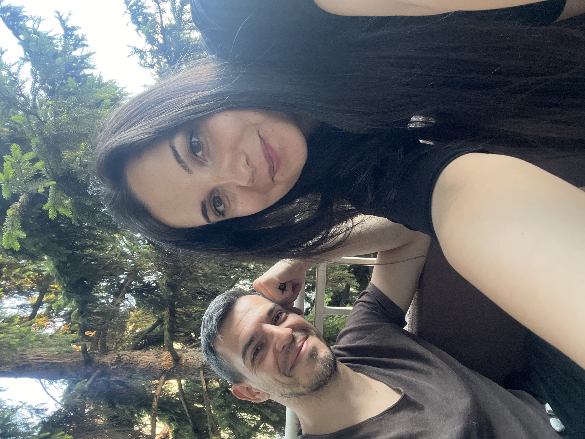 A selfie taken outdoors in Sofia, Bulgaria, features a man and a woman. The man, on the left, has short, graying hair and a beard, and wears a brown t-shirt. He leans his head on his hand, resting his elbow on a railing, and smiles at the camera. The woman, on the right, has long, dark hair and wears a black t-shirt. She also smiles at the camera. They are positioned in front of a large evergreen tree, with a railing visible in the background. The lighting suggests it is daytime.