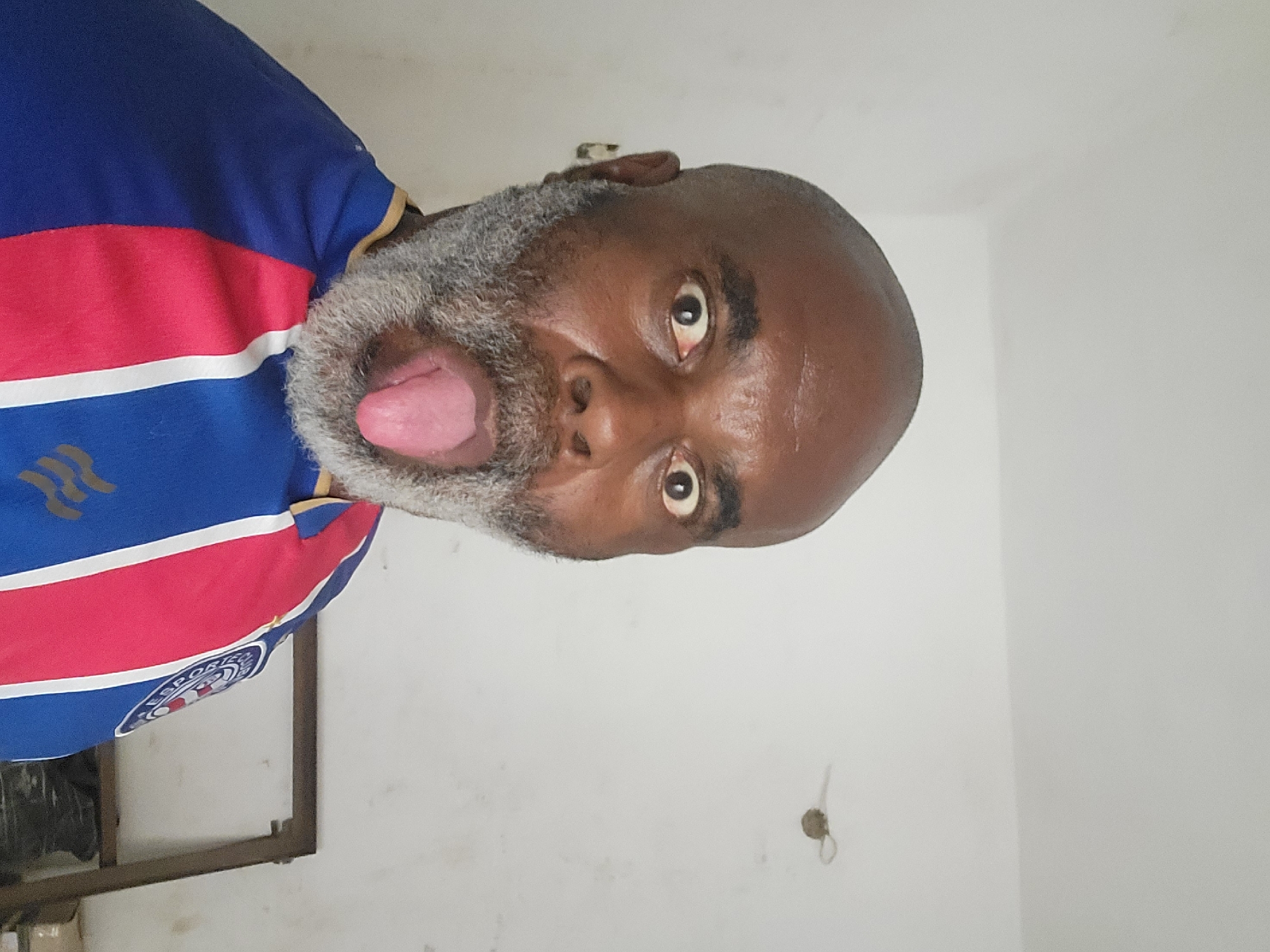A bald, middle-aged Black man with a gray beard and mustache is making a face with his tongue sticking out. He is wearing a blue shirt with red and white stripes and the logo of Esporte Clube Bahia. He is indoors, with white walls behind him. No location data is provided to confirm it is from the city of Maiburiji, Nigeria.