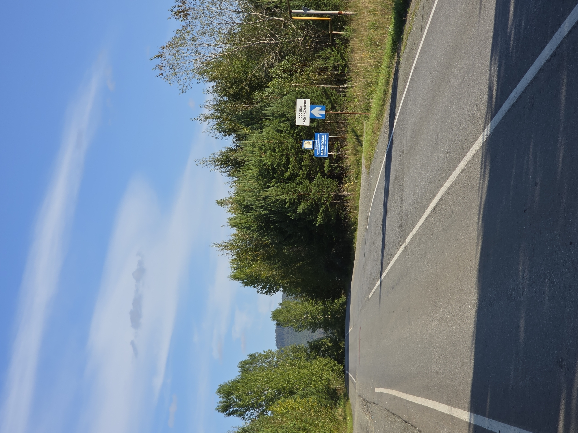 The image depicts a two-lane asphalt road extending into a natural landscape under a partly cloudy blue sky. The road is bordered by a grassy verge on the right and a gravel shoulder on the left. A dense line of green trees, primarily coniferous and deciduous, forms a backdrop to the road, with a distant mountain range visible behind them.
On the right side of the road, several signs are placed near the tree line. One white sign with blue text and an arrow indicates "ООО ПКФ ПРОМИНДУСТРИЯ" (OOO PKF PROMINDUSTRIA) and points forward with a blue arrow. Below this, a smaller blue sign displays a symbol and the text "Кусинский муниципальный район МАГНИТСКОЕ" (Kusinsky Municipal District Magnitskoe), also with an arrow pointing forward. A yellow pole is also visible to the right of these signs. The overall impression is a rural or semi-rural setting on a clear day, likely a highway leading into or out of the mentioned administrative district in Russia, given the presence of Cyrillic text on the signs. No people are visible in the image.