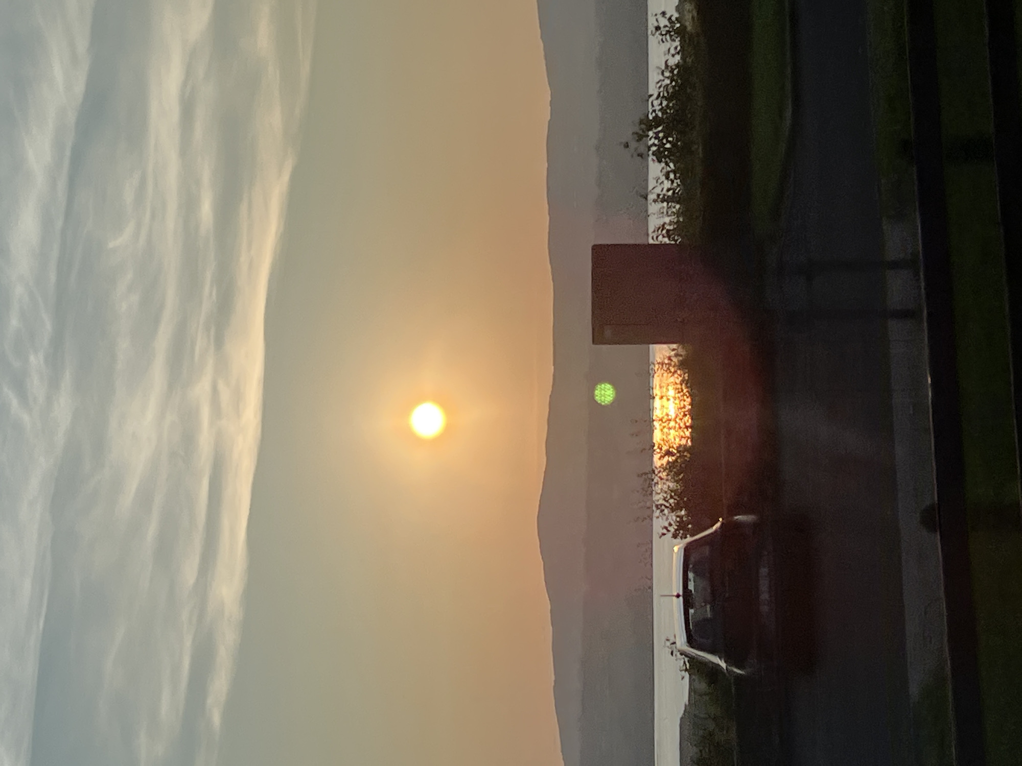The image depicts a scenic sunset over a body of water, likely Lough Swilly, as viewed from Letterkenny, Ireland. The sun, a bright orb, is positioned in the upper center of the frame, casting a warm, orange glow across the sky and reflecting on the water. Silhouetted against the sunset are rolling hills in the distance. In the foreground, a dark-colored car is parked on a paved surface. To the right of the car, a rectangular structure, possibly a bin or a small building, stands near a patch of vegetation. The overall scene is serene, with the soft light of dusk creating a tranquil atmosphere.