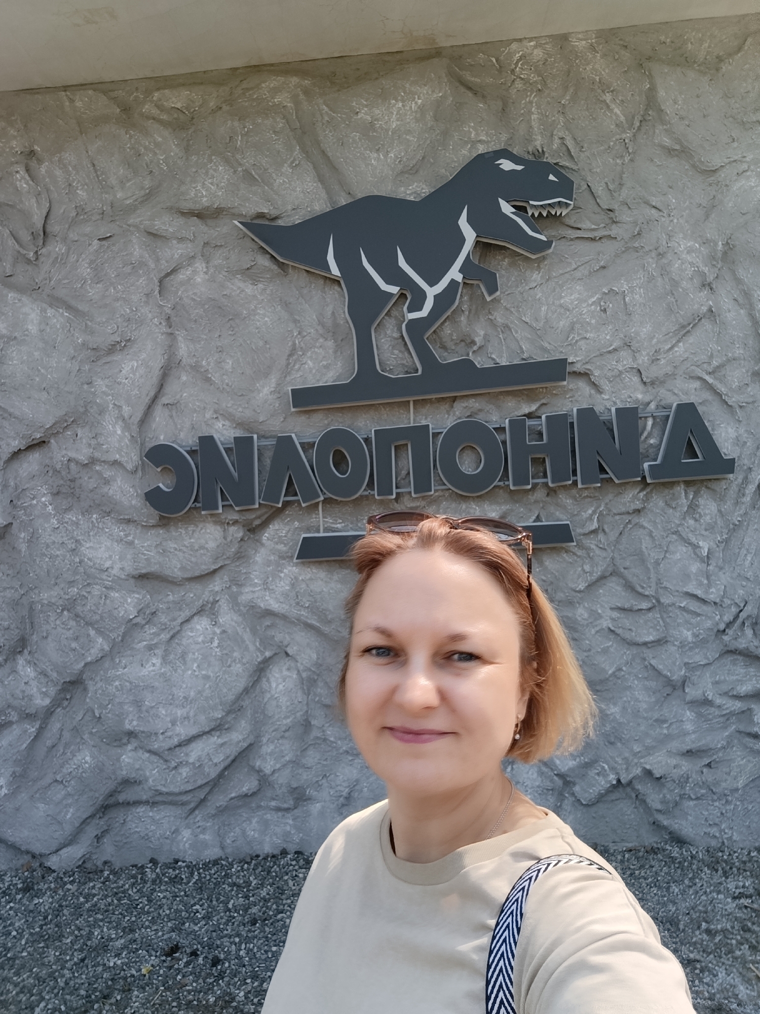 A woman is standing in front of a sign with a dinosaur at the Izhevsk Zoo in Izhevsk, Russia. The sign is gray and mounted on a wall with a rough, stone-like texture. The dinosaur is a Tyrannosaurus Rex with white accents. The name of the zoo is written below the dinosaur in Cyrillic. The woman is wearing a beige t-shirt and has sunglasses on her head. A shoulder strap is visible on her left shoulder. The ground in front of the wall appears to be covered with small gray pebbles.