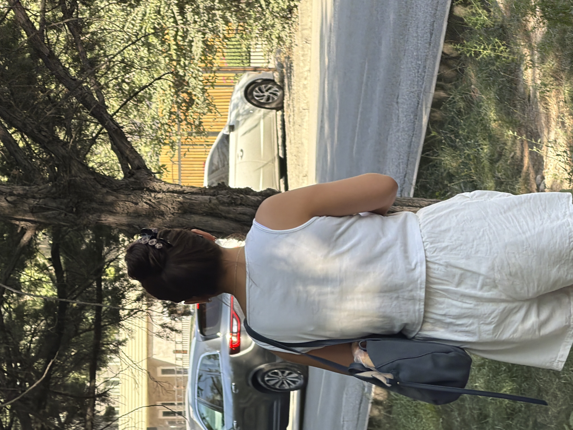 A woman stands with her back to the camera, facing a tree trunk and a street with two cars. She wears a white tank top and white shorts, and carries a dark blue shoulder bag. Her hair is pulled up in a bun with a decorative clip. In the background, a silver SUV is parked on the left, and a white sedan is parked on the right. The scene appears to be outdoors, possibly in a residential area, with trees and vegetation visible.