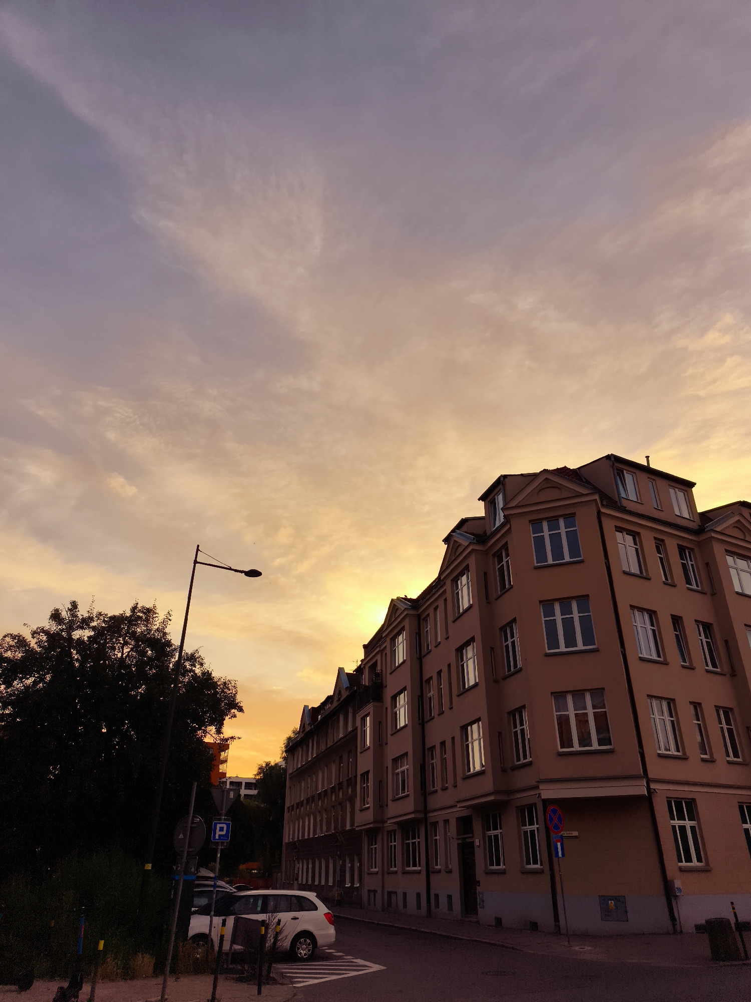 The image shows a street corner in Gdańsk, Poland, at sunset. A tall, multi-story building with light brown walls and white-framed windows dominates the right side of the frame. The building has a mansard roof. To the left of the building, a dark green tree is silhouetted against the sky, and a street lamp with a bent pole stands in front of it. A white station wagon is parked on the street. The sky is filled with wispy clouds that are illuminated by the setting sun, creating a gradient of orange, yellow, and purple hues. Several street signs are visible, including a blue "P" sign for parking. The ground is paved with asphalt.