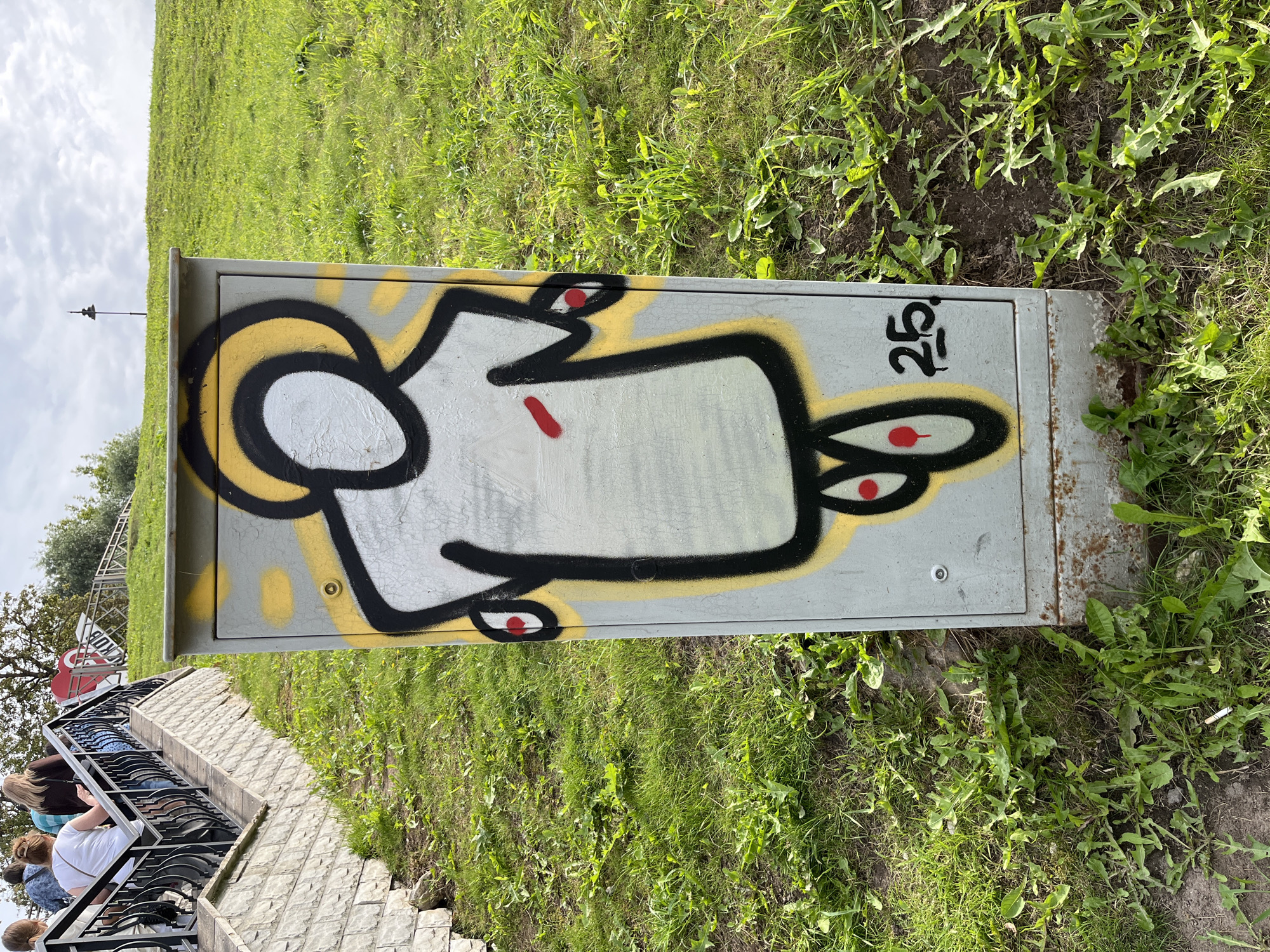 An outdoor, eye-level, medium shot captures a graffiti-adorned utility box set against a grassy hillside. The box itself is light gray and shows signs of wear, with some rust at the bottom. Dominating the front of the box is a stylized, monochrome graffiti figure with a halo. The figure has a black outline with yellow accents and red dots for eyes and a red mark on its chest. To the right of the figure, the number "25" is spray-painted in black.
The surrounding environment is a gently sloping green lawn, with patches of dirt and scattered weeds. In the background, a stone staircase with black metal railings ascends the hill. Several individuals, seen from behind, are walking up the stairs. Further up the hill, a red sign with white lettering that reads "ПСКОВ" (Pskov) is visible, along with a lamppost on the horizon. The sky is overcast with scattered clouds. The overall scene appears to be a public outdoor space in Pskov, Russia.