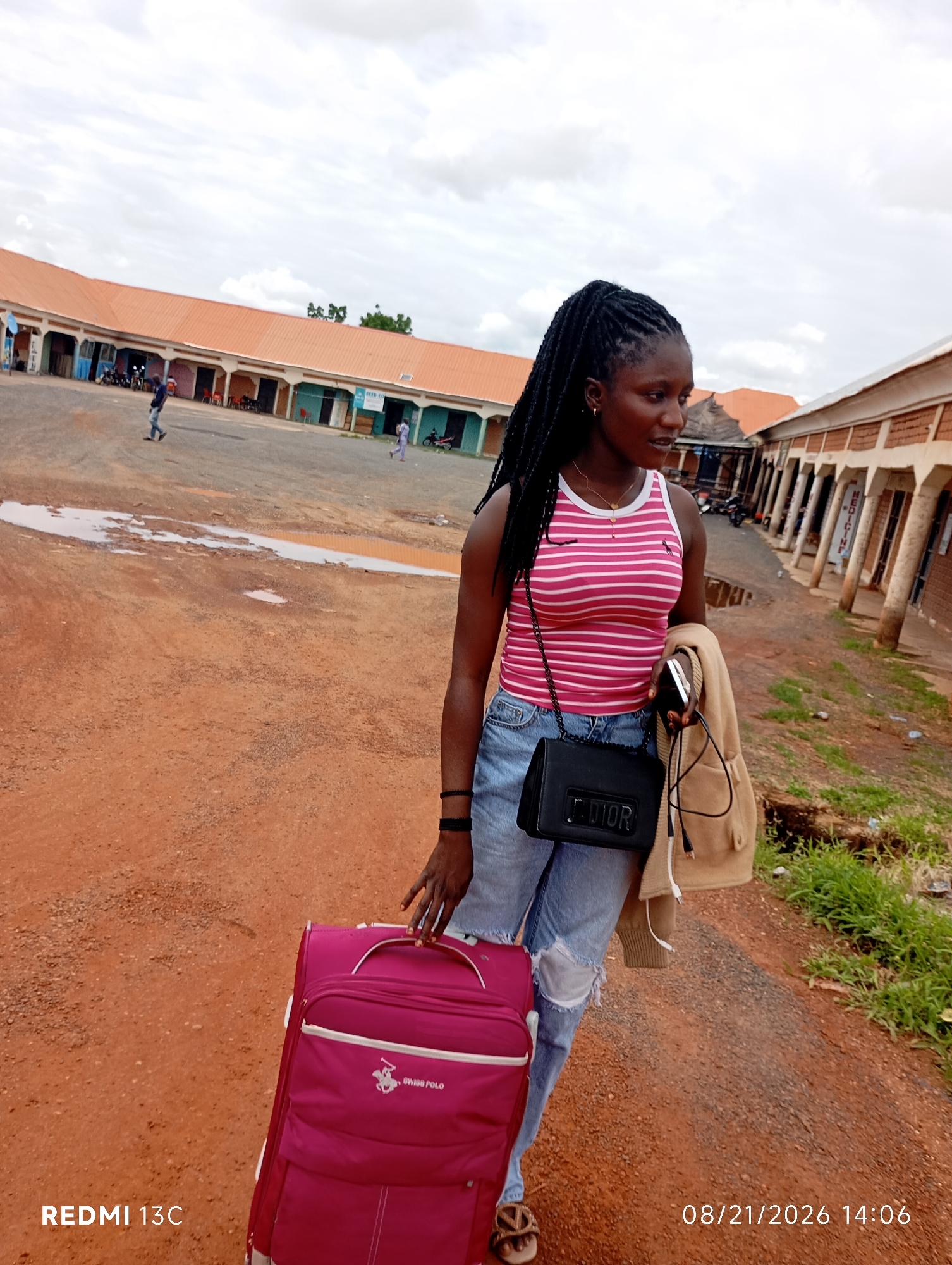 In the city of Shendam, Nigeria, a young Black woman with braided hair stands outdoors, holding a pink suitcase and a beige jacket draped over her arm. She wears a pink and white striped tank top, ripped blue jeans, and sandals. A black Dior purse is slung across her body. The ground is reddish-brown dirt, with a few puddles of water and some sparse green grass. In the background, there are buildings with orange roofs and covered walkways, typical of a market or administrative area. Several people are visible in the distance, some walking and others near motorcycles. The sky is overcast. The timestamp "08/21/2026 14:06" is visible in the lower right corner, and "REDMI 13C" is in the lower left.