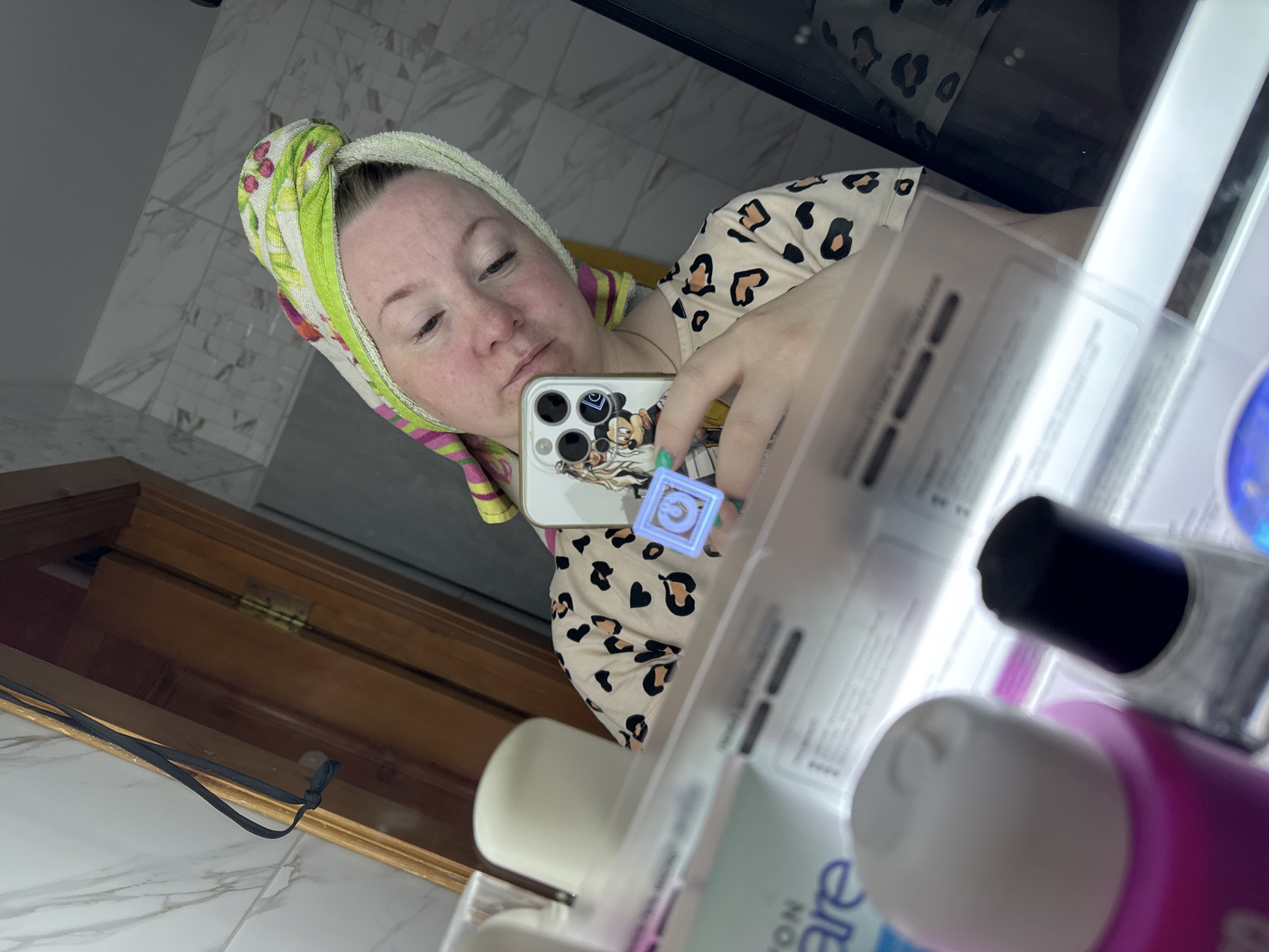 The image depicts a person with fair skin and reddish hair wearing a towel wrapped around their head, taking a selfie in a bathroom. The person is wearing a leopard-print shirt. They are holding a phone with a case featuring cartoon characters. The bathroom has white marble tile on the walls and floor. A wooden cabinet with a black drawstring is partially visible in the lower left corner of the frame. Reflected in the bathroom mirror are various toiletries including tubes of lotions/creams and a nail polish bottle. The image was likely taken in a residential bathroom. The location in the city of Chaikovsky, Russia is not discernible from the image.