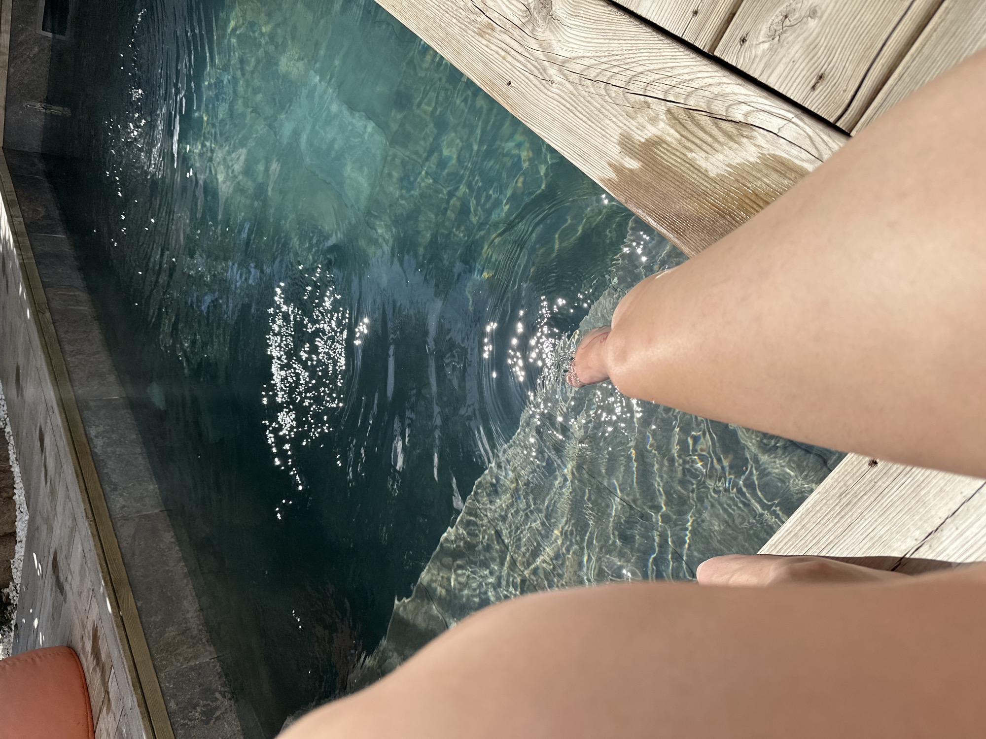 A medium, angled shot depicts a partially visible individual dipping their foot into a pool of water. The setting includes wooden decking to the right and a stone-lined pool on the left, filled with clear water reflecting sunlight. The person's leg and foot are positioned towards the water's edge, creating ripples. The composition suggests an intimate, personal moment by the pool. It is plausible to assume the location is somewhere near La Couarde-sur-Mer, France given the user input.