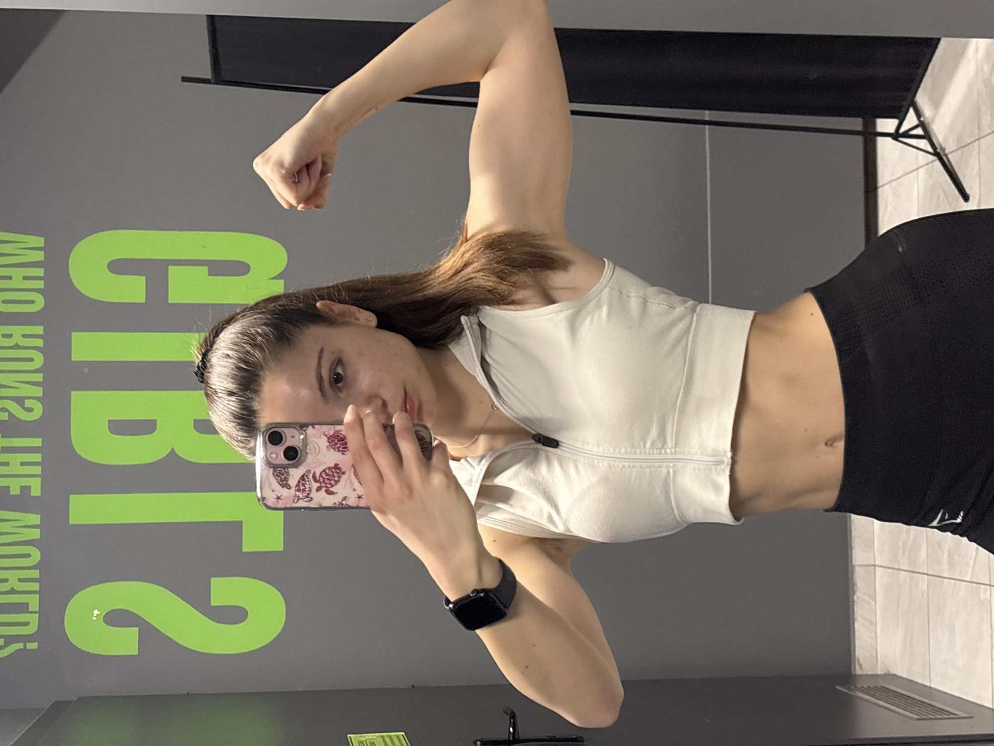 A young woman is posing for a selfie in front of a mirror in a gym, likely in Varna, Bulgaria. She is wearing a white sports bra and black leggings. Her left arm is flexed, showing off her bicep. She is holding a smartphone in her right hand, taking the picture. The background features a large, green inscription on the wall, reading "GI2 BT2". A black exercise equipment stand is partially visible in the upper right corner of the image. The gym appears to have grey walls and tiled flooring. The woman is also wearing a smartwatch.