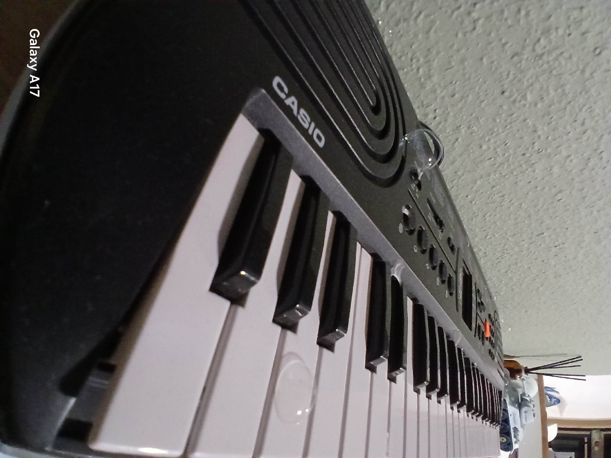 An upward-angled view captures a black Casio electronic keyboard positioned indoors in Yekaterinburg, Russia, on what appears to be a tabletop or similar surface next to a textured, light-colored wall. The keyboard occupies the majority of the frame, extending diagonally from the lower-left foreground towards the upper-right background. The white "CASIO" logo is clearly visible in the lower-left on the keyboard's black frame.
The keyboard features black and white keys, with several white keys prominently displayed in the foreground and mid-ground. A notable detail is a small, clear water droplet resting on one of the white keys in the mid-right portion of the image. Another, smaller droplet is visible on the black control panel to the left, near a textured, ridged design element. The control panel also displays various black circular buttons and a distinctive orange rectangular button in the upper-mid section.
In the background, on the right side of the image, several objects are discernible. These include a small, dark-colored vase holding several thin, dark sticks, likely part of an incense or reed diffuser set. To the right and slightly behind the vase, a light-colored toy tank, possibly a military model, is visible. The wall behind these objects and extending across the left side of the frame has a rough, stucco-like texture. The bottom-left corner of the image includes a "Galaxy A17" watermark, indicating the device used to capture the photograph. No people are present in the scene.
