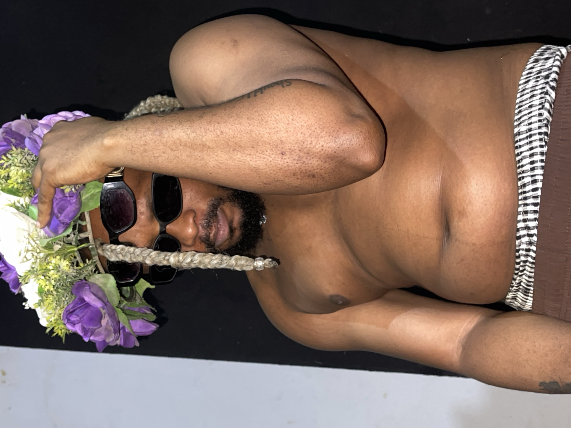 A shirtless man with brown skin, a beard, and blonde braids is wearing sunglasses and a floral headdress. He is standing with his right arm raised, touching the headdress. He is wearing brown shorts with a black and white striped waistband. The background is a dark, solid color with a vertical white and gray section on the left side. The lighting suggests an indoor setting. The image is undated and no specific location in Jos, Nigeria, is evident from the visual information alone.