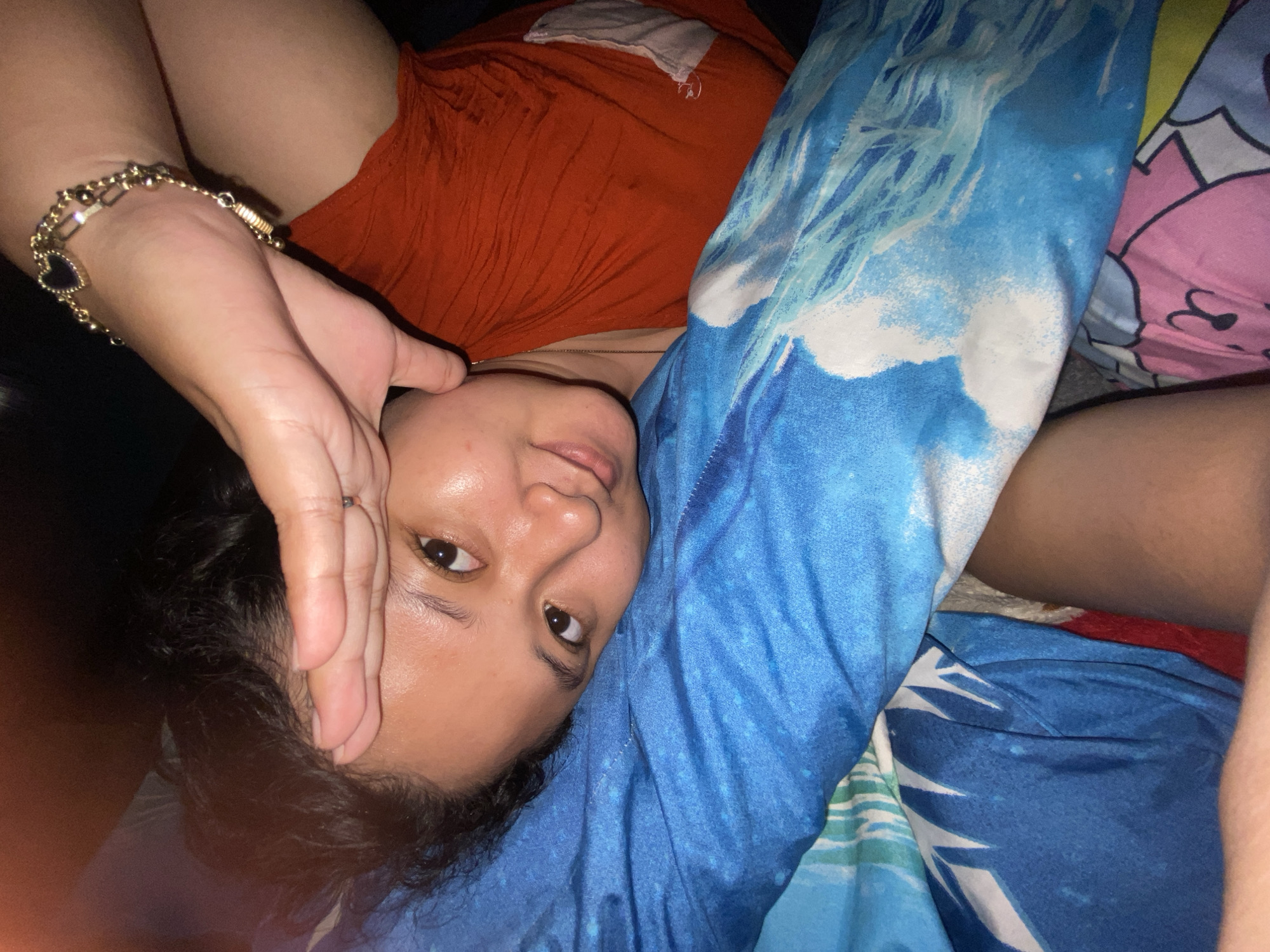 A person is lying on a bed, with a blue patterned pillow underneath their head and a pink patterned pillow partially visible on the right. The person is wearing an orange shirt. The left hand is raised, with fingers near their face. A gold bracelet is on the left wrist. The environment appears to be indoors, dimly lit. The location is assumed to be Gelam, Indonesia, based on a previous user prompt, but cannot be confirmed from the image itself.