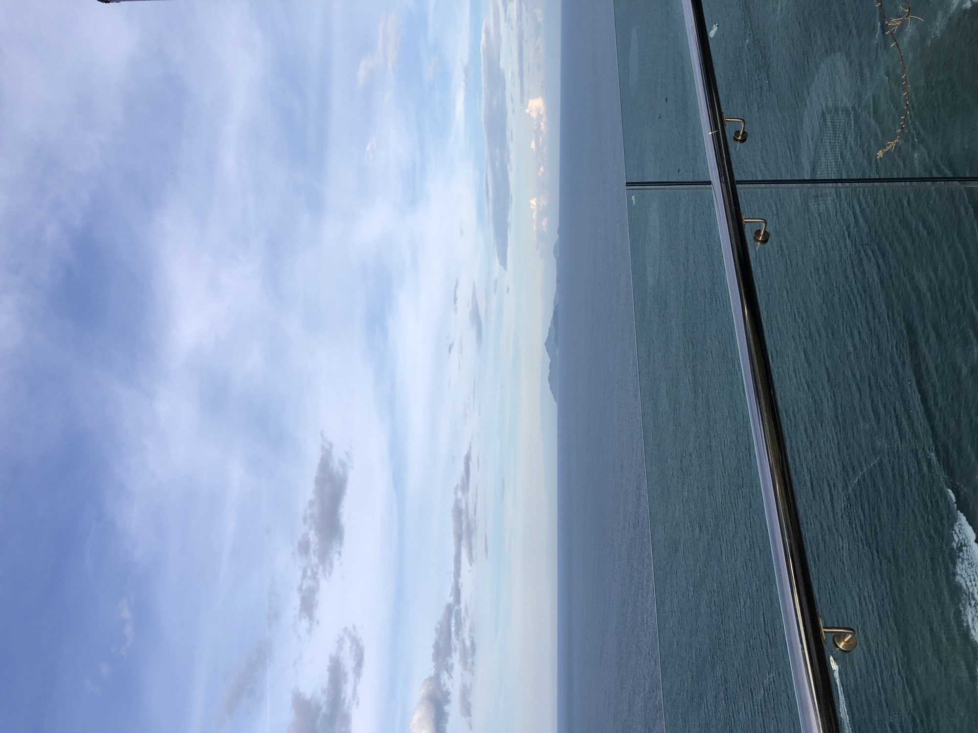 The image depicts a panoramic view of the sea from a high vantage point, likely a balcony or rooftop in Đà Nẵng, Vietnam. In the foreground, a polished chrome handrail with glass panels forms a barrier, suggesting it's part of a modern building. The sea, a deep teal color, stretches to the horizon, dotted with distant, hazy islands or landmasses that appear mountainous. The sky is a pale blue, streaked with wispy white clouds. The overall impression is one of a serene, coastal landscape under a partly cloudy sky.
