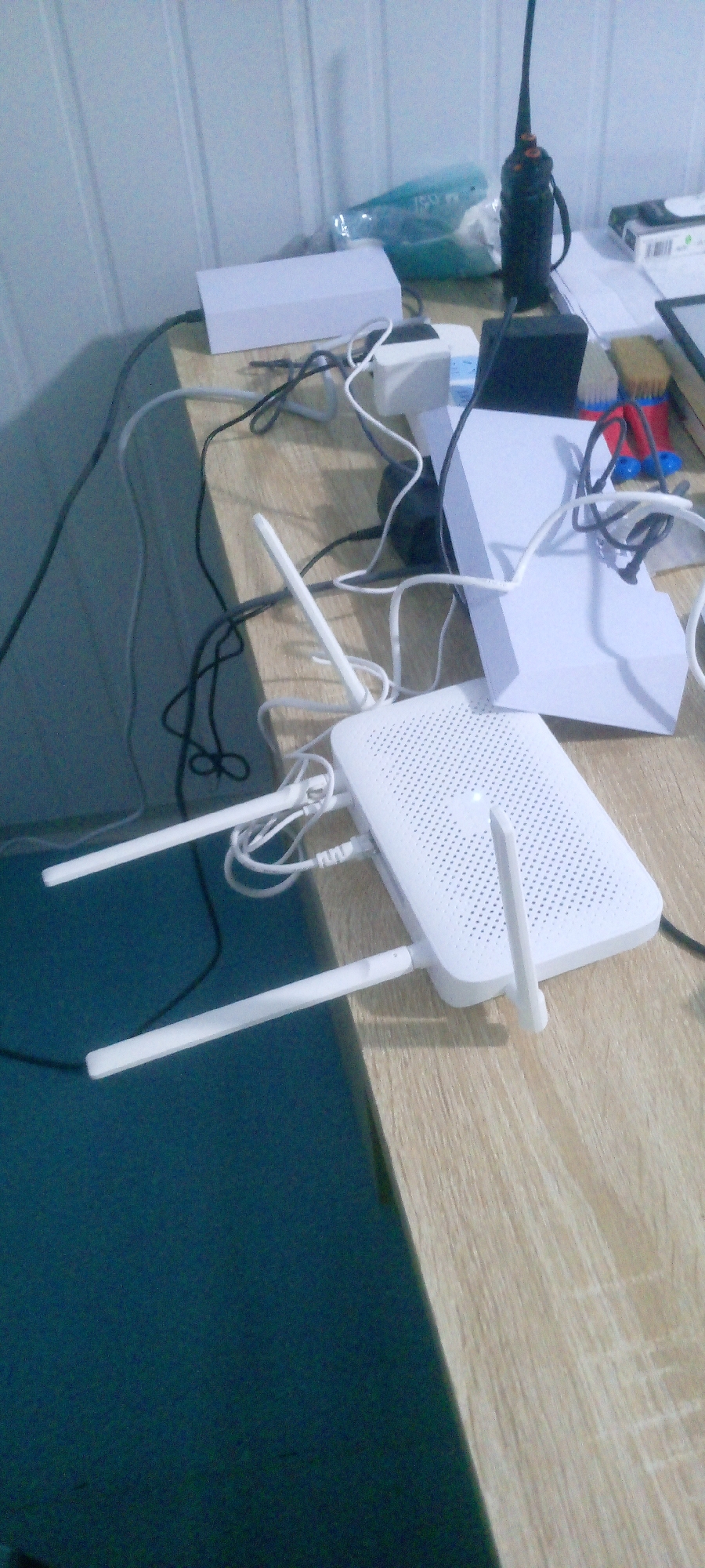 The image depicts a cluttered desk with various electronic devices and cables, in a room with light-colored, paneled walls. The primary focus is a white Wi-Fi router with two antennas, positioned on the right side of the desk, connected to multiple cables. Behind the router, there's a white box-like object, a black power adapter, and a small black walkie-talkie. To the left, a white rectangular box is visible, along with more cables. A green plastic bottle and a pack of cigarettes are also on the desk. The surface of the desk is a light brown wood grain. The bottom left of the image shows a dark teal surface, possibly a mat or a desk pad. There are no people visible in the image. The scene appears to be an indoor setting, likely a home office or workspace, but its specific location in Indo, Nigeria cannot be determined from the visual information provided.