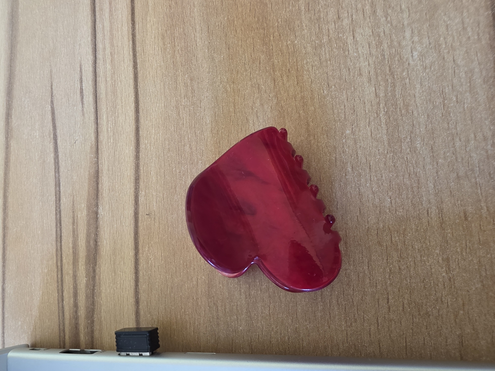 A glossy, red, heart-shaped hair clip lies on a light brown, wooden surface. The clip is positioned slightly to the right of the center of the image, with its pointed end facing left. To the left of the clip, a silver electronic device is visible, with a small black USB adapter protruding from its side. The wooden surface has a visible grain pattern, with horizontal lines running across the frame. The lighting appears to be natural, casting a soft illumination on the scene. There are no people present in the image, and no actions or interactions are depicted. The overall scene is static and appears to be a close-up shot of desk accessories.